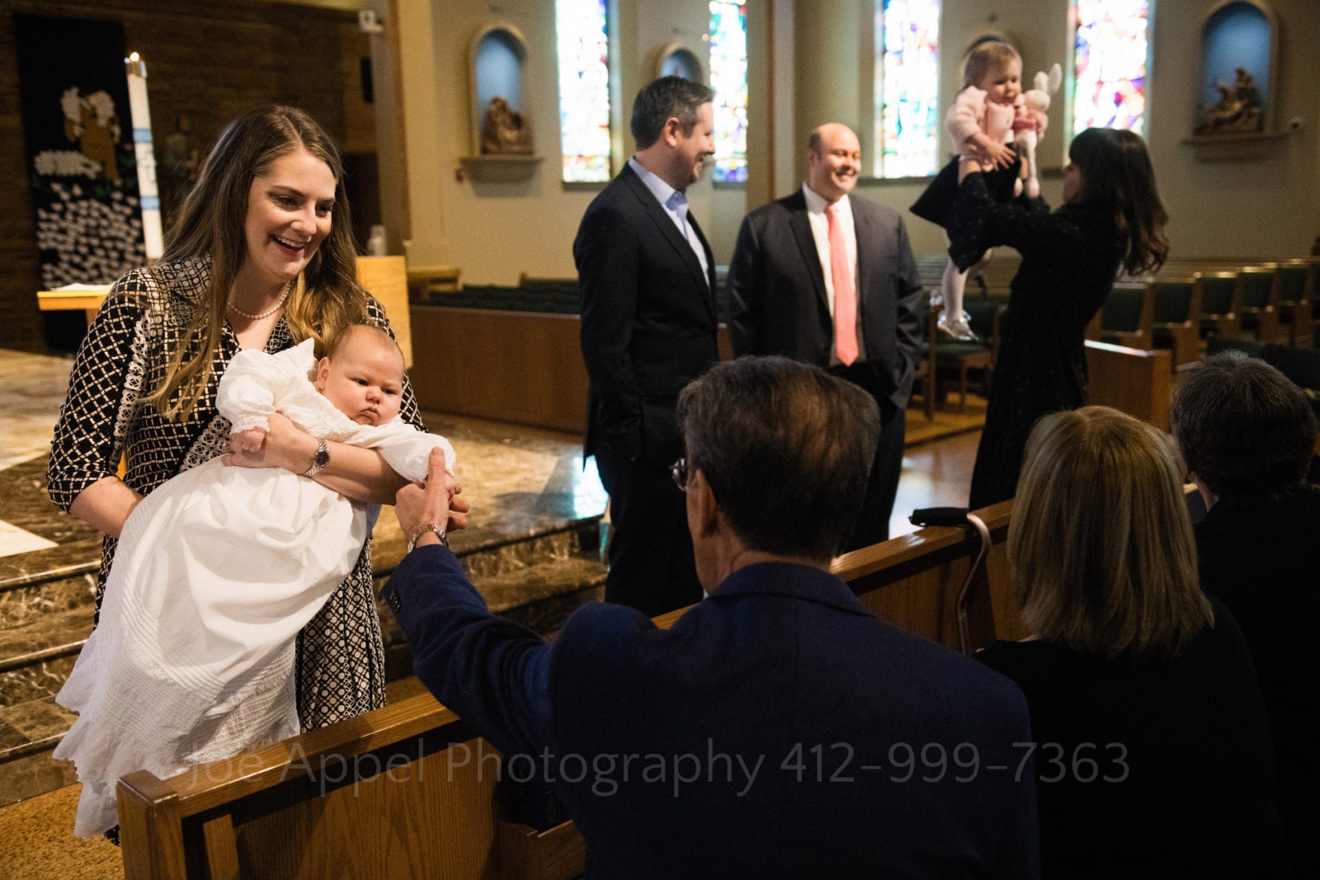 A woman holds a baby in front of an older man who reaches out to touch the child's hand. In the background three adults stand with an older child. A woman lifts the child into the air during a Baptism at Saints John and Paul Parish.