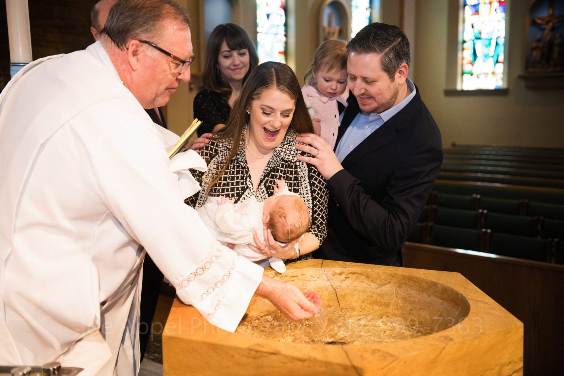 During a Baptism at Saints John and Paul Parish a deacon splashes water on to the head of a baby held over a baptismal font by his smiling mother. The father holds the couple's other child and has his hand on the mother's shoulder. Another woman (the godmother) in the background smiles as she watches.