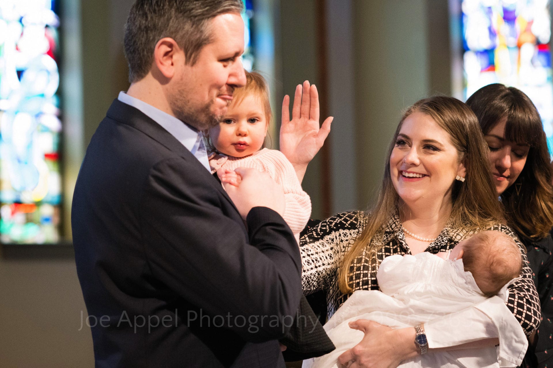 A mother holding a baby in one arm holds the other hand up in a blessing to her husband who holds the couple's older child during a Baptism at Saints John and Paul Parish.