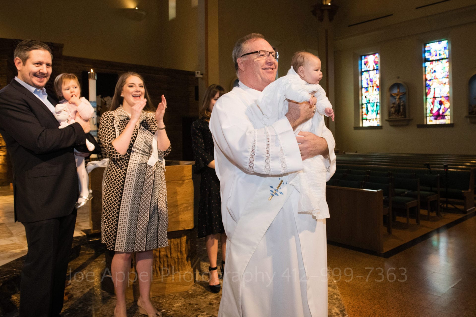A deacon holds up a newly baptized baby for the congregation to see during a Baptism at Saints John and Paul Parish. The mother and father are standing behind him applauding.