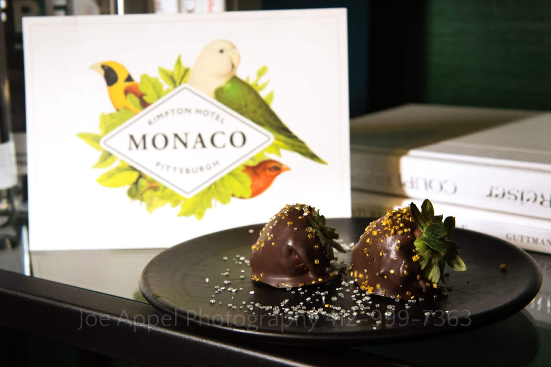 two chocolate covered strawberries with gold sprinkles sit on a black plate in front of a card from the hotel monaco in pittsburgh 