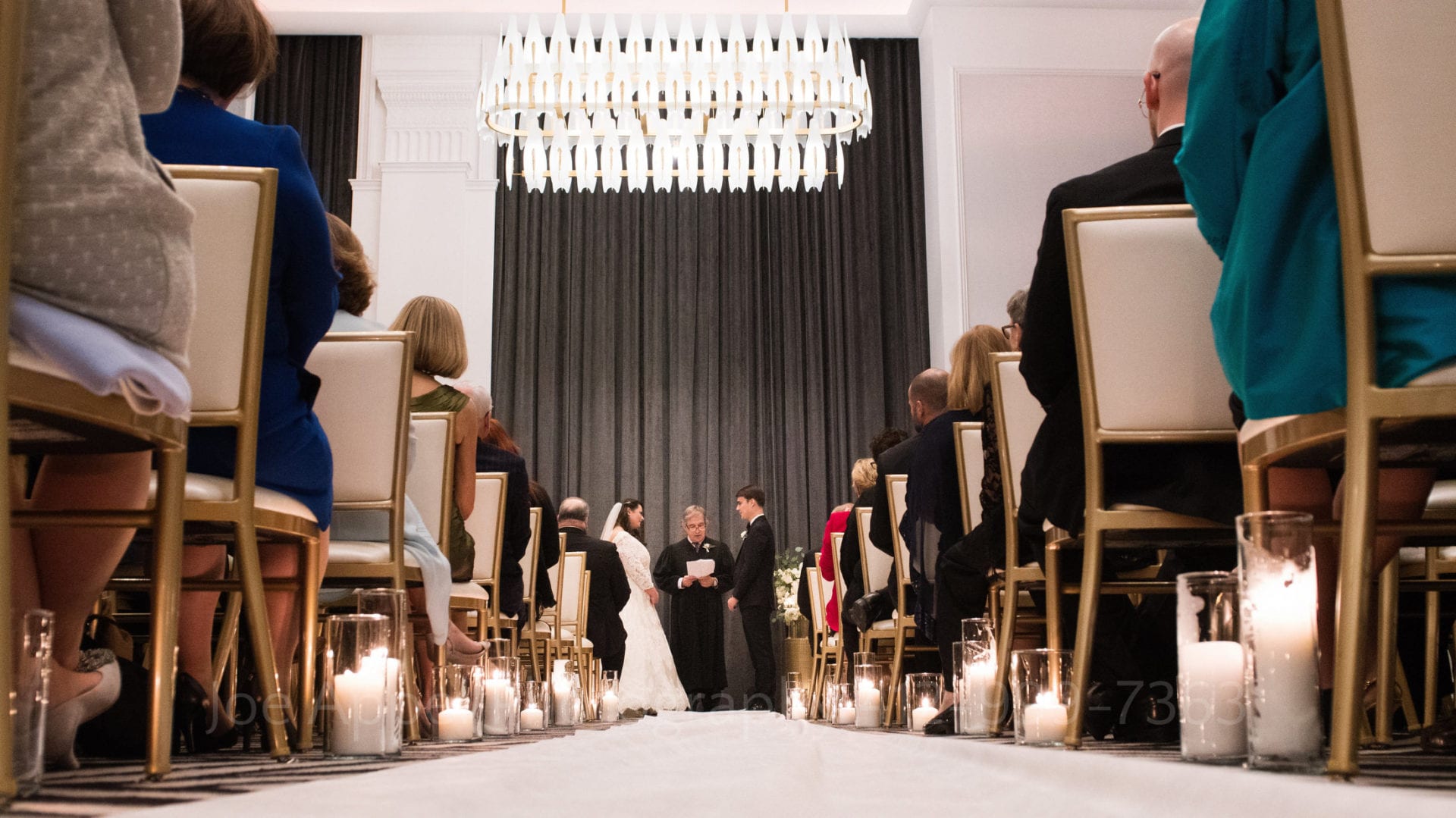 guests sit along an aisle decorated with candles as they watch a bride and groom exchange vows