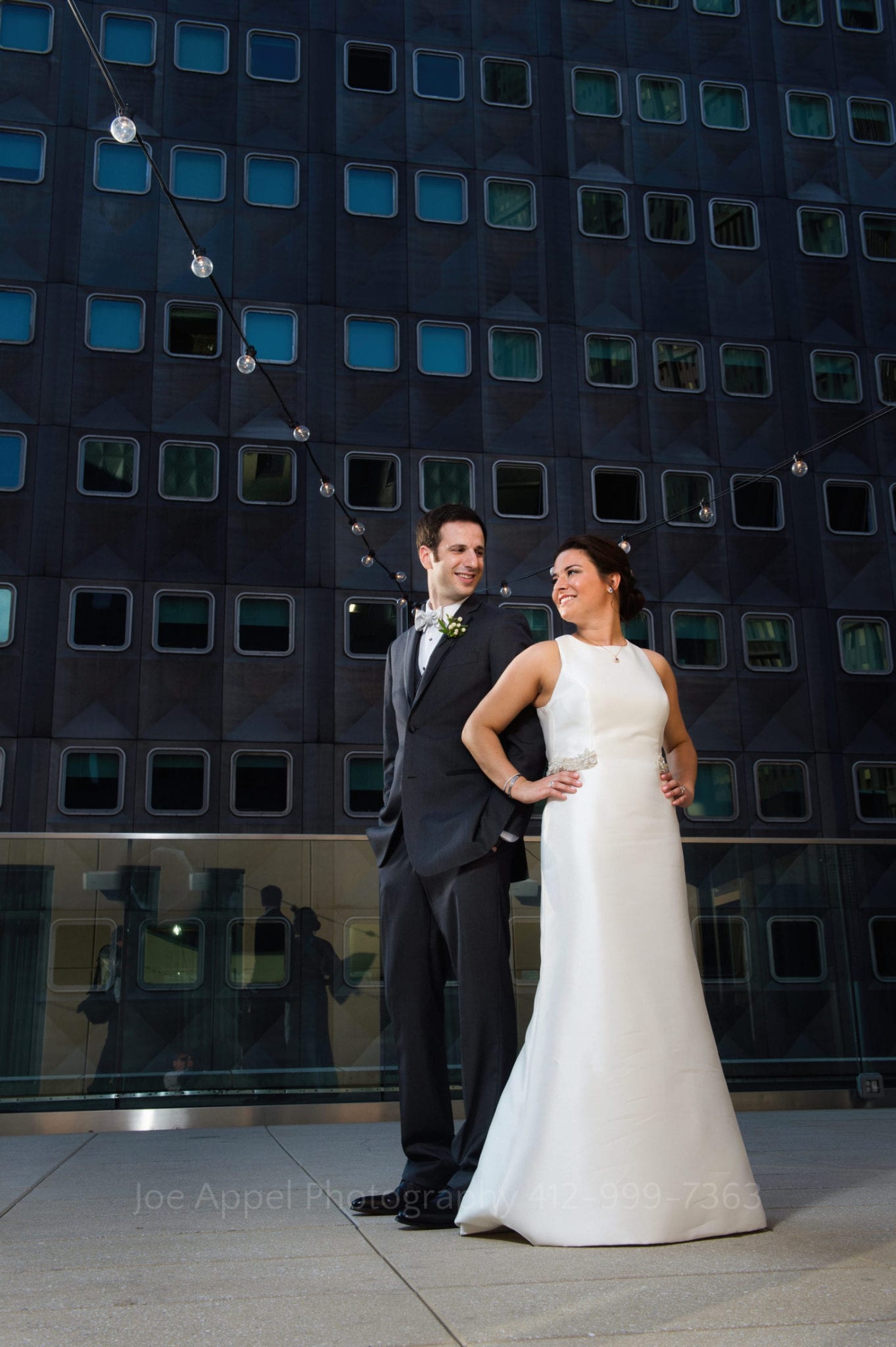 a bride and groom stand in front of a black building and strings of lights