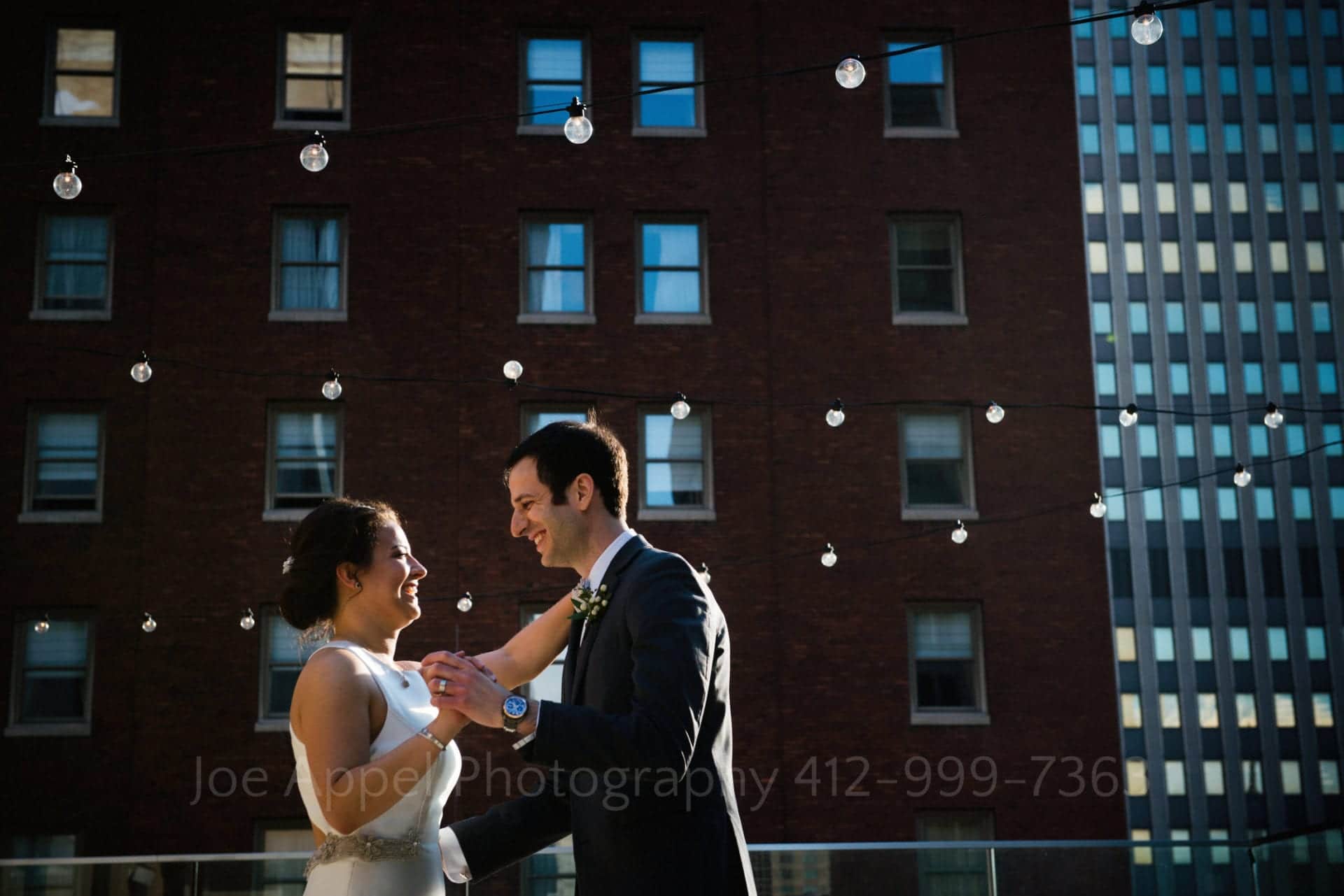a bride and groom dance in front of city buildings and strings of lights