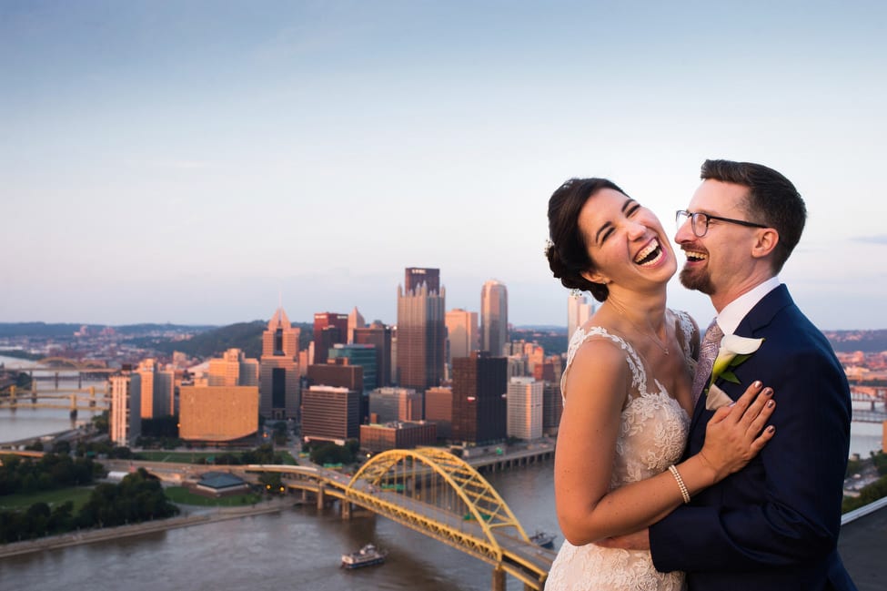 A laughing bride and groom embrace as they stand on Mt. Washington with the skyline of downtown Pittsburgh behind them.