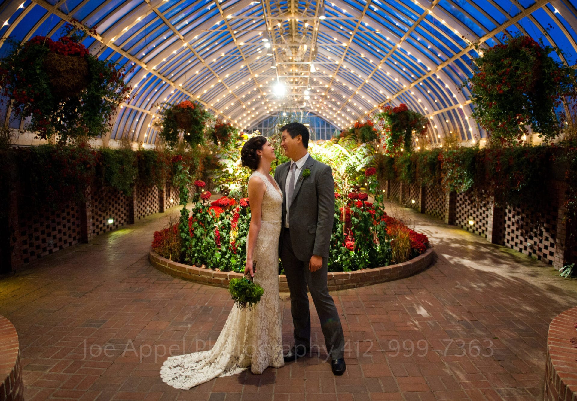 a bride and groom smile at each other in front of a display of red flowers Phipps Conservatory Weddings