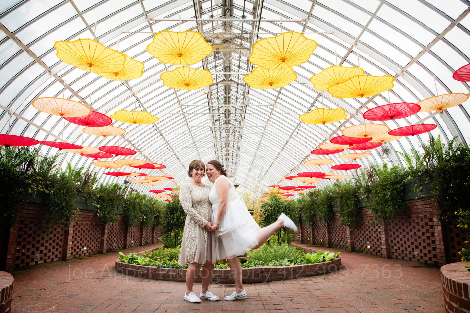 two brides wearing dresses and converse stand under red orange and yellow umbrellas Phipps Conservatory Weddings