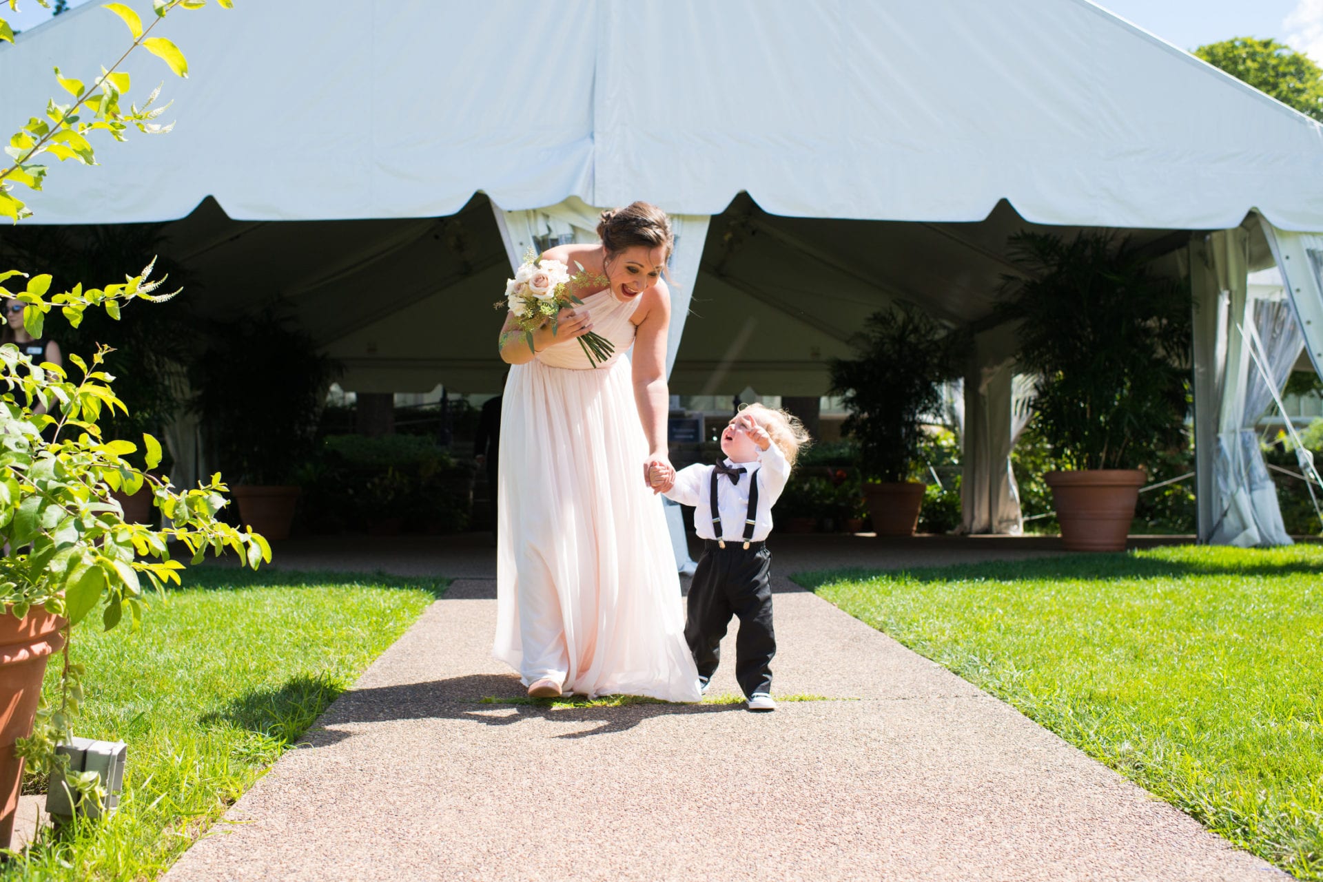 a bridesmaid in a pink dress walks with a little boy wearing black suspenders and a bow tie Phipps Conservatory Weddings