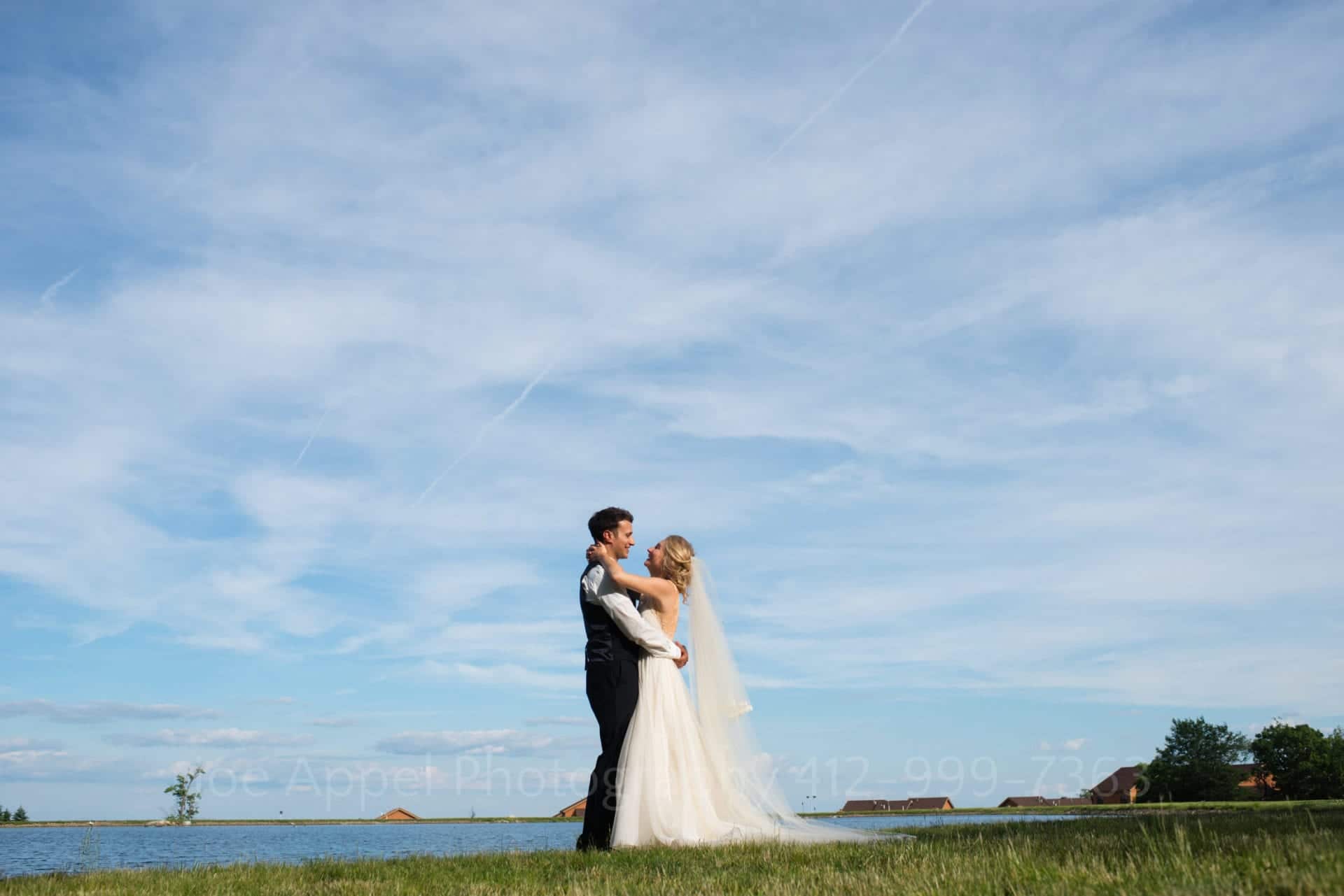 a bride and groom embrace in front of a lake and a blue sky Seven Springs Weddings