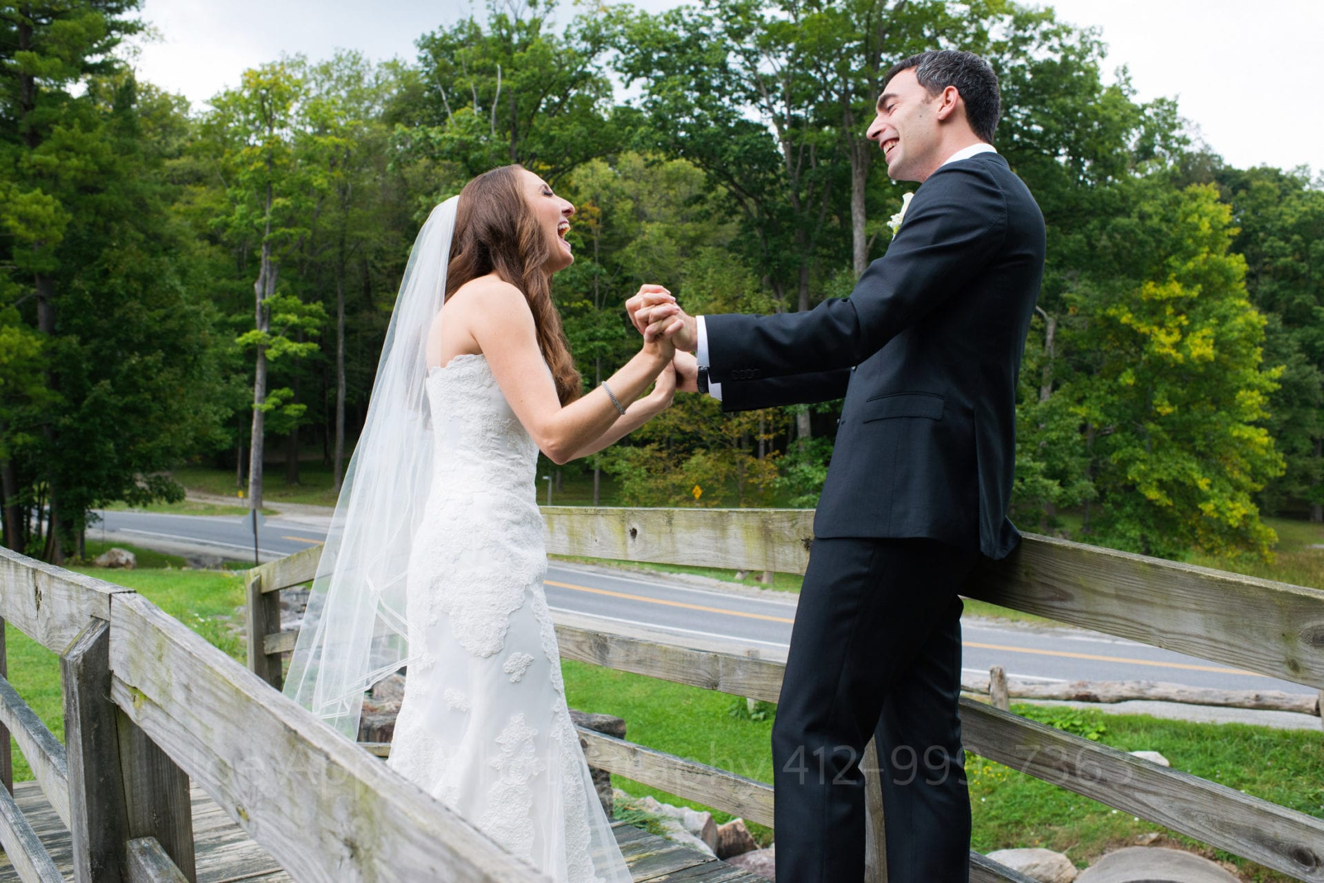 A bride and groom hold each others hands in front of them as they stand on a small bridge that crosses a stream. They're expressions are joyful.