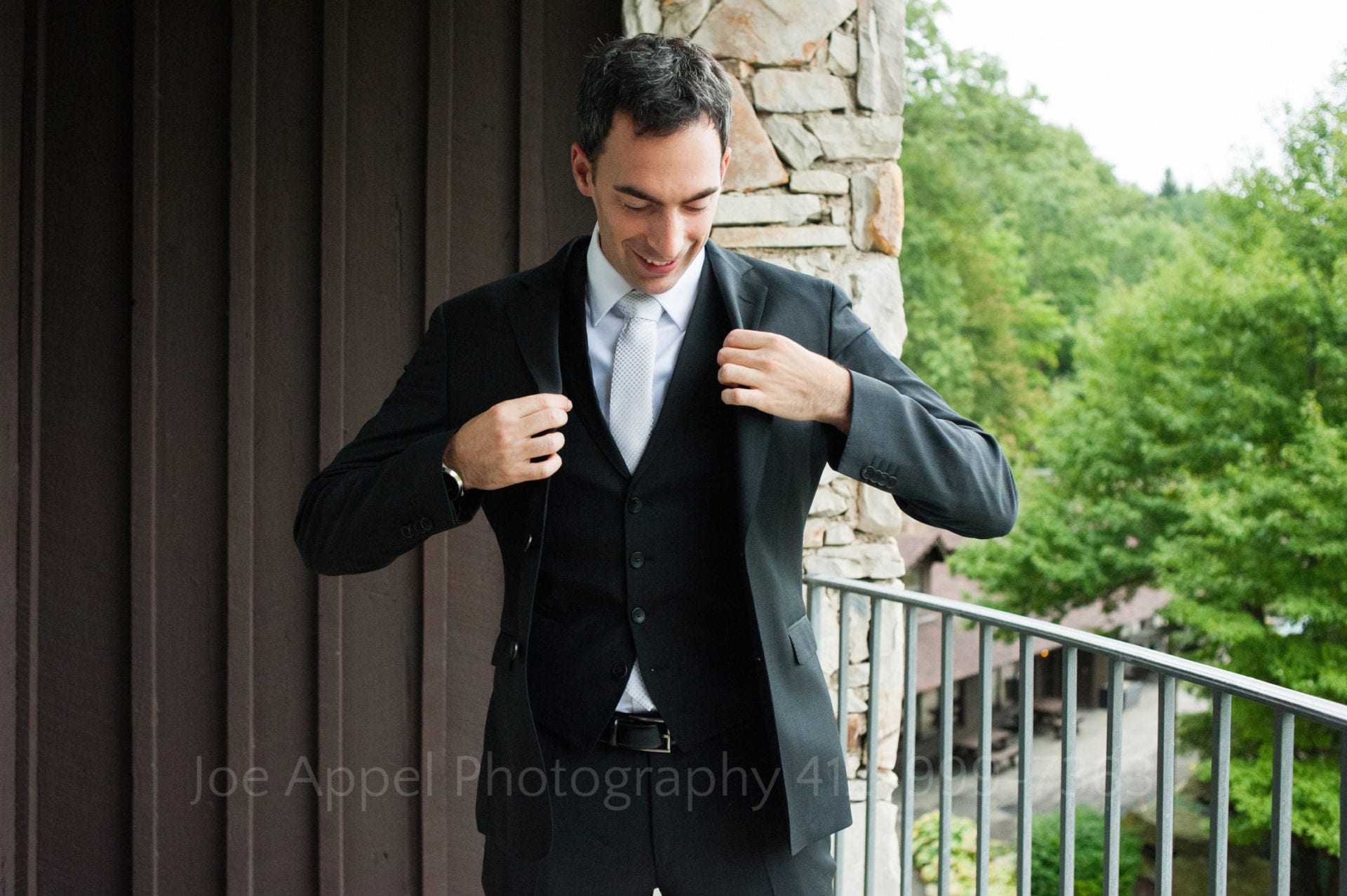A groom puts on his jacket as he stands on a balcony made of stone and wood.