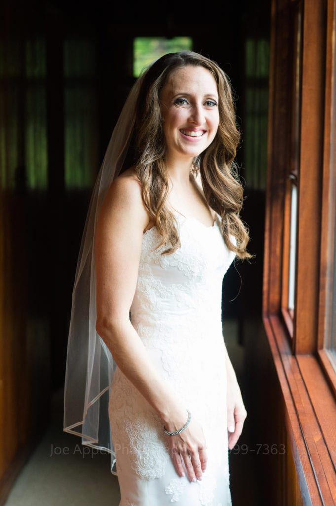 A smiling bride wearing a white dress and veil stands by a window before her Seven Springs Resort Wedding.