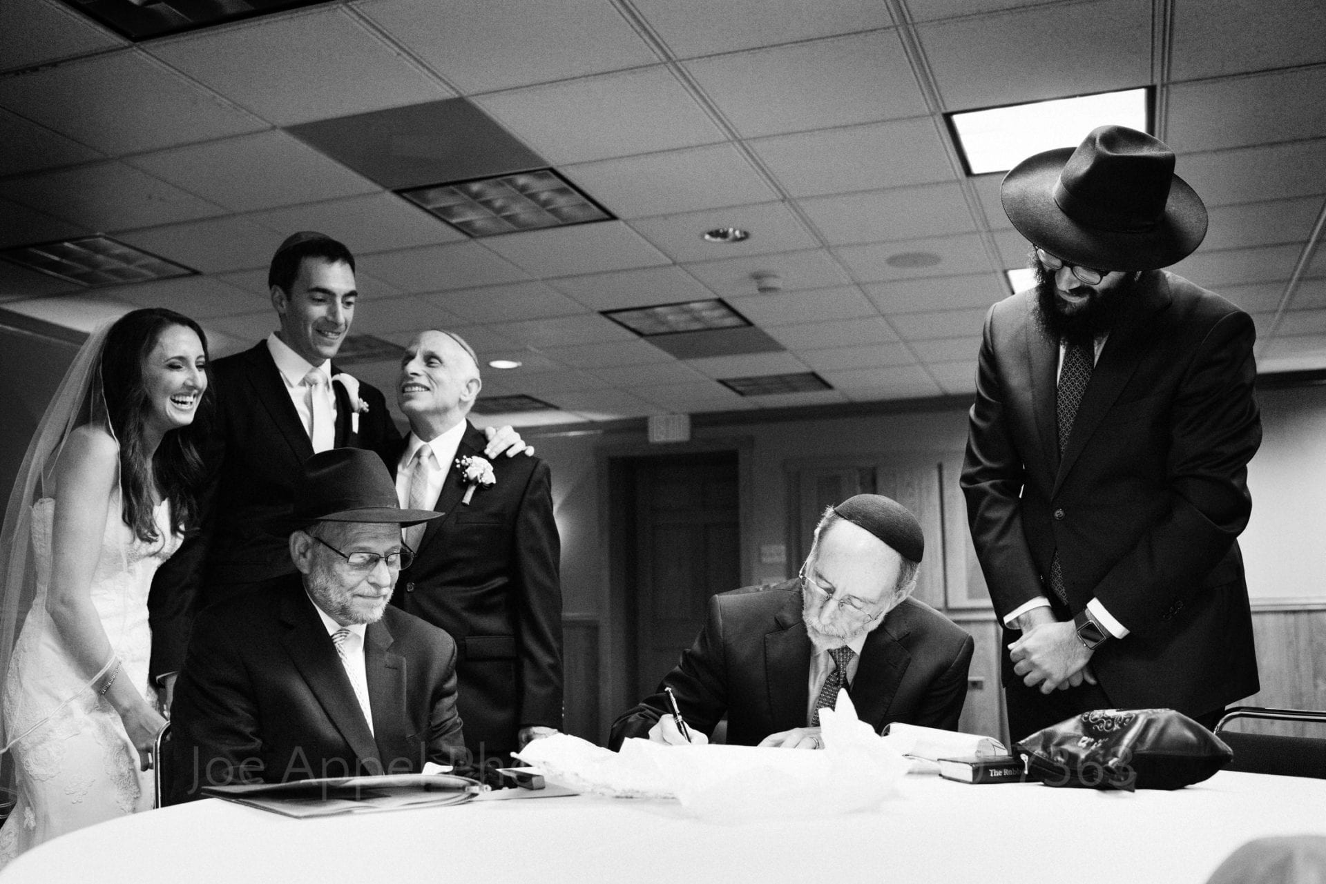 Rabbis seated at a table sign paperwork for a couple during their marriage ceremony. The couple stands behind them along with the bride's father.