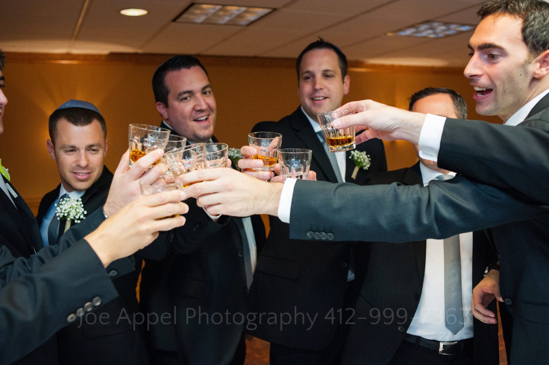 A group of men in black suits touch glasses of whiskey in the middle of their circle.