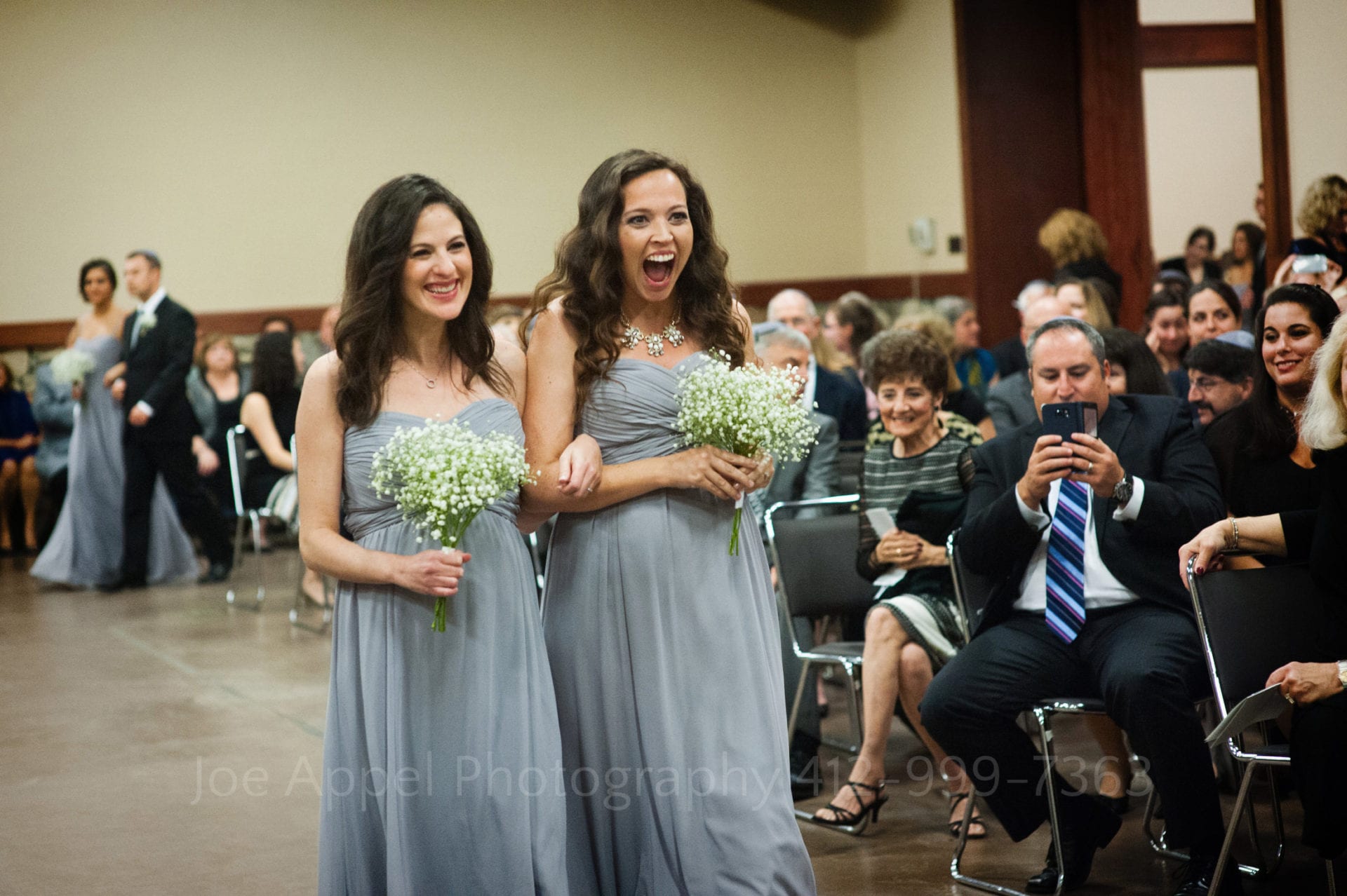 Two pregnant smiling bridesmaids carry flowers and walk down an aisle at a wedding.