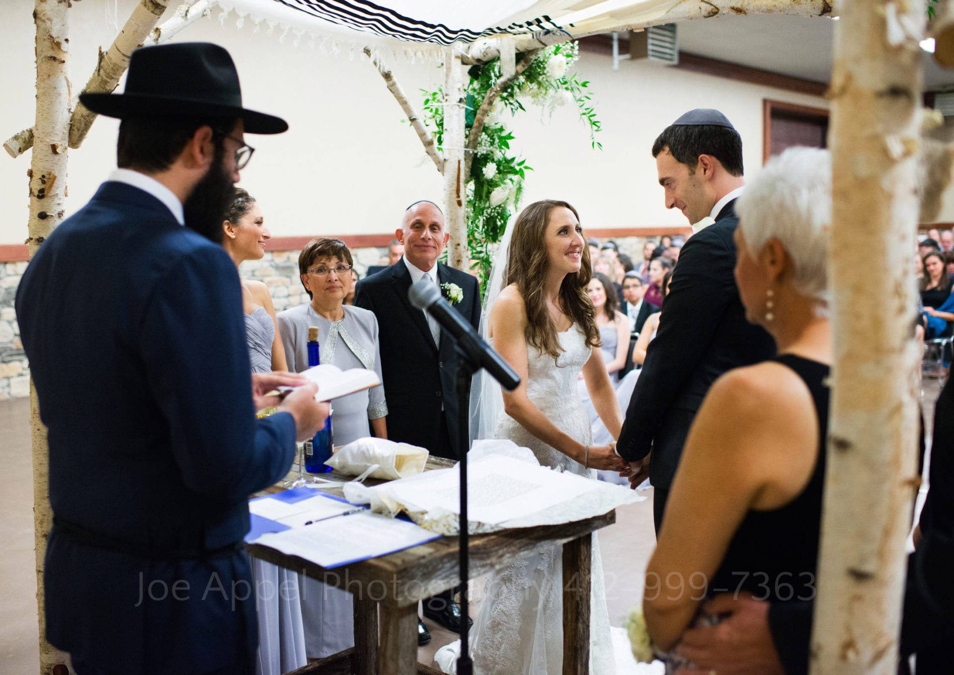 Seen from behind the rabbi, a couple exchanges wedding vows beneath a chuppah.