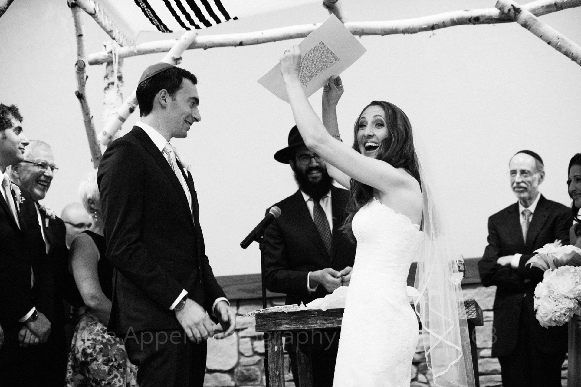 A smiling bride holds a ketubah above her head and looks at the guests during her wedding ceremony.