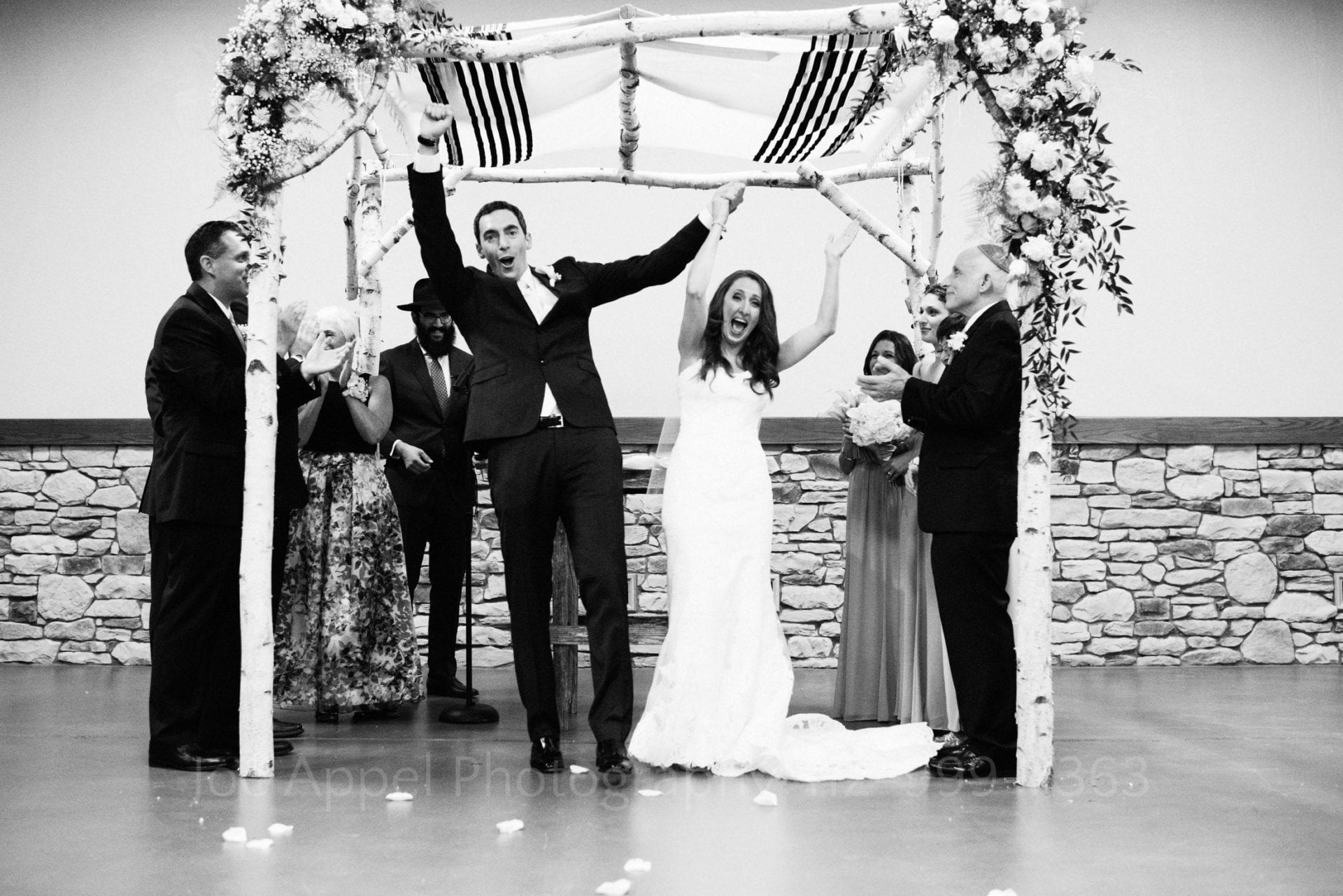 A bride and groom cheer and hold their hands in the air beneath the chuppah at the end of their wedding ceremony.