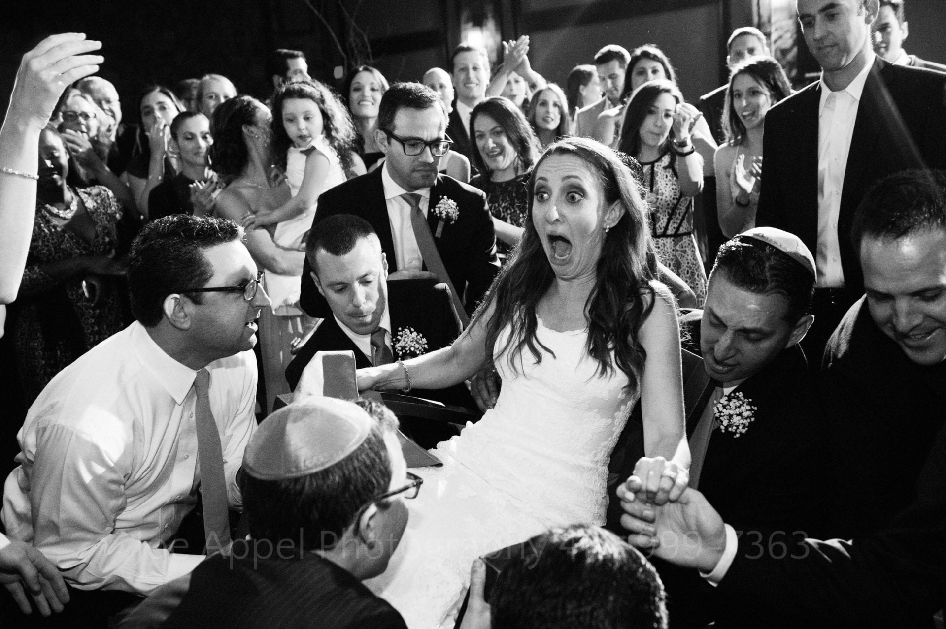 A bride has a look of shocked surprise on her face as she is lifted in a chair by a group of men.