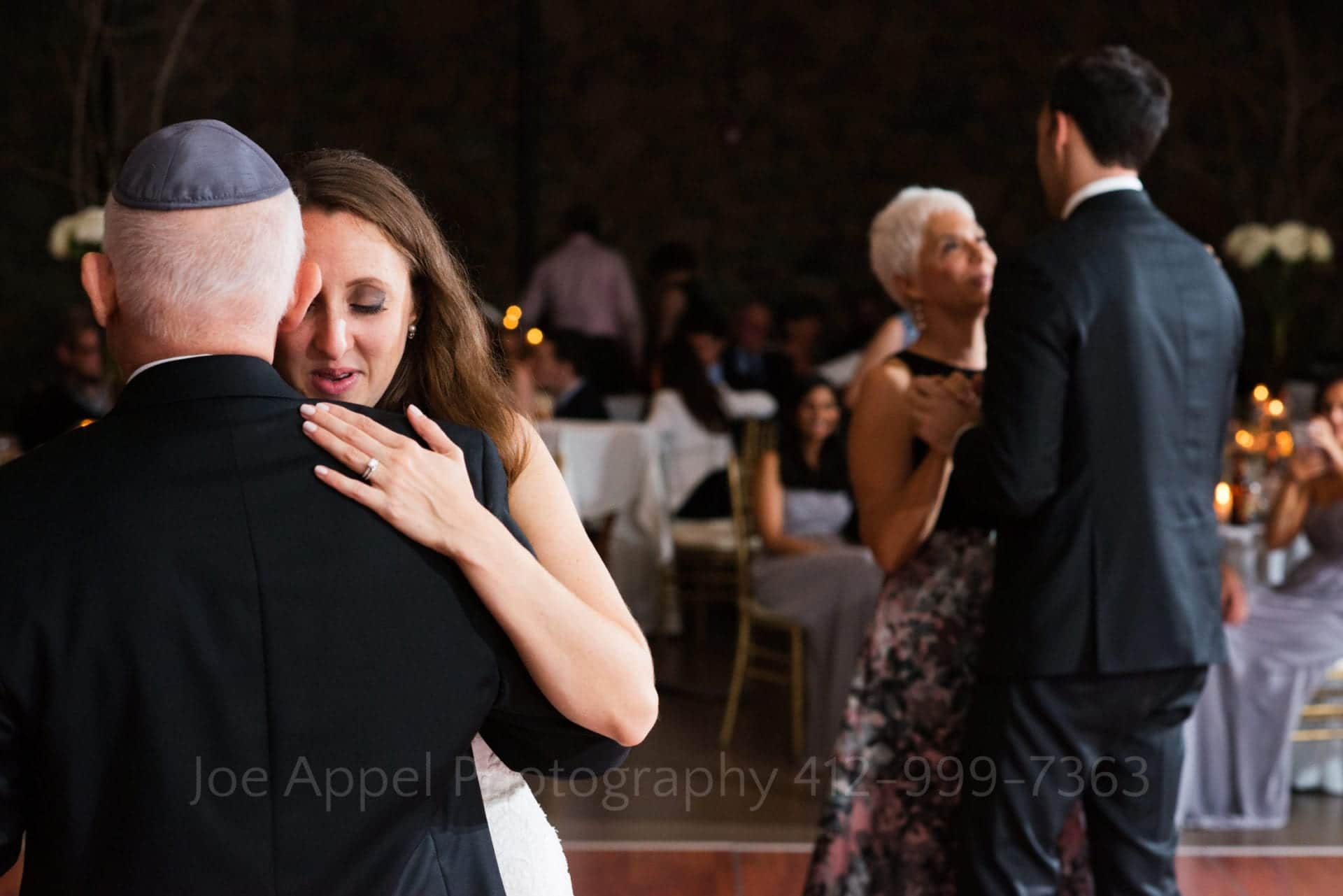 A bride holds on to her father as they dance in the foreground while the groom and his mother dance in the background.