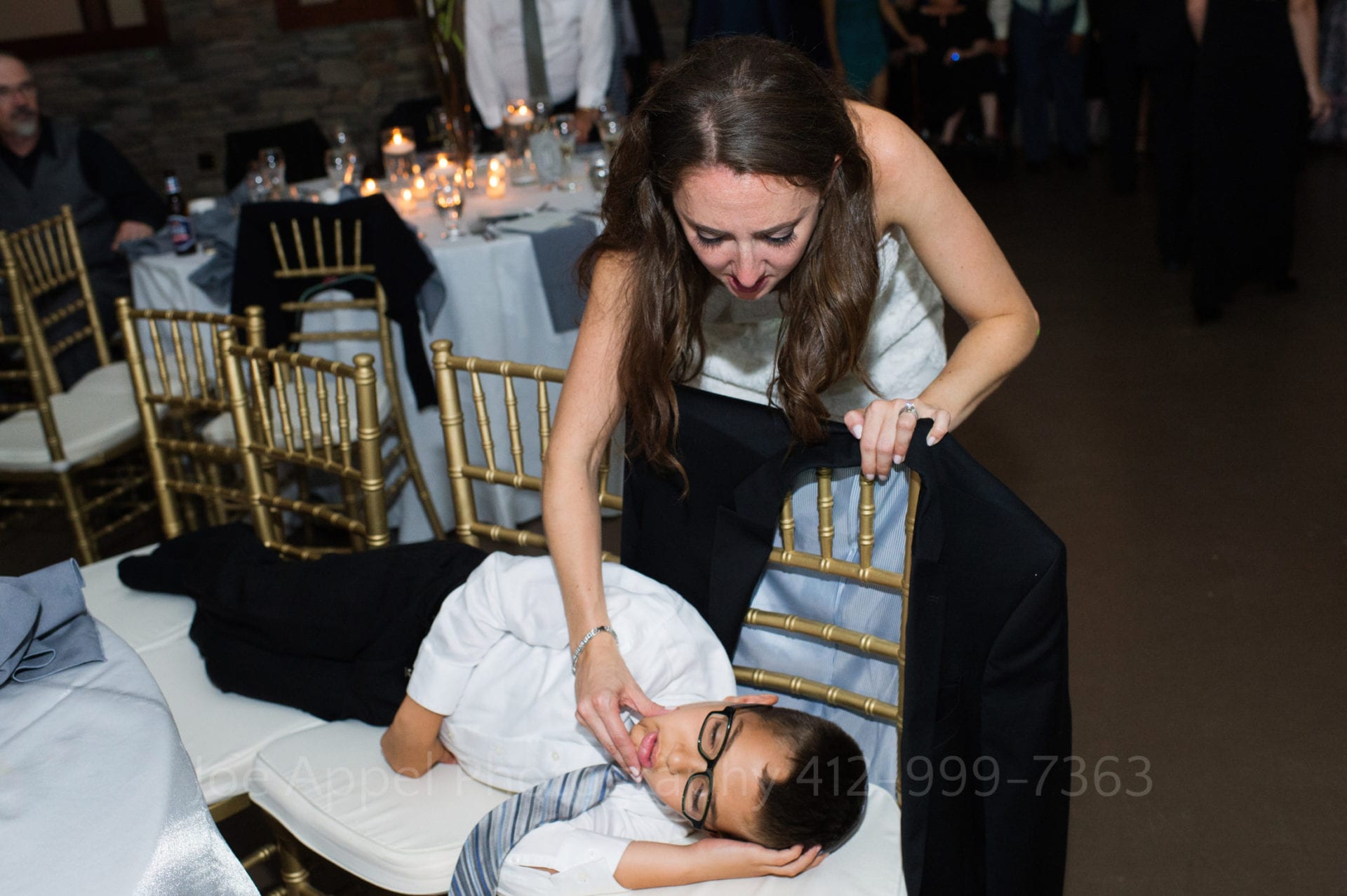 A bride reaches down to touch a child who is sleeping on several chairs that have been pushed together.