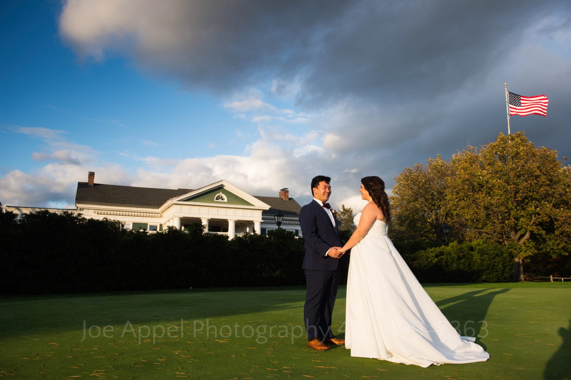 A bride in a white dress stands facing her groom wearing a dark blue suit, brown shoes, and a bow tie. There is a green and white mansion behind them with a hedgerow between them. The skies are blue with dark, puffy clouds. An American flag flies in the upper right part of the frame. Pittsburgh Golf Club Weddings.