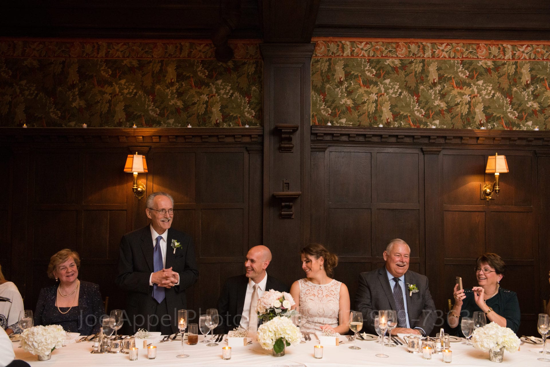 a bride and groom sit at the dinner table and smile as their family gives toasts during their Pittsburgh Golf Club Weddings