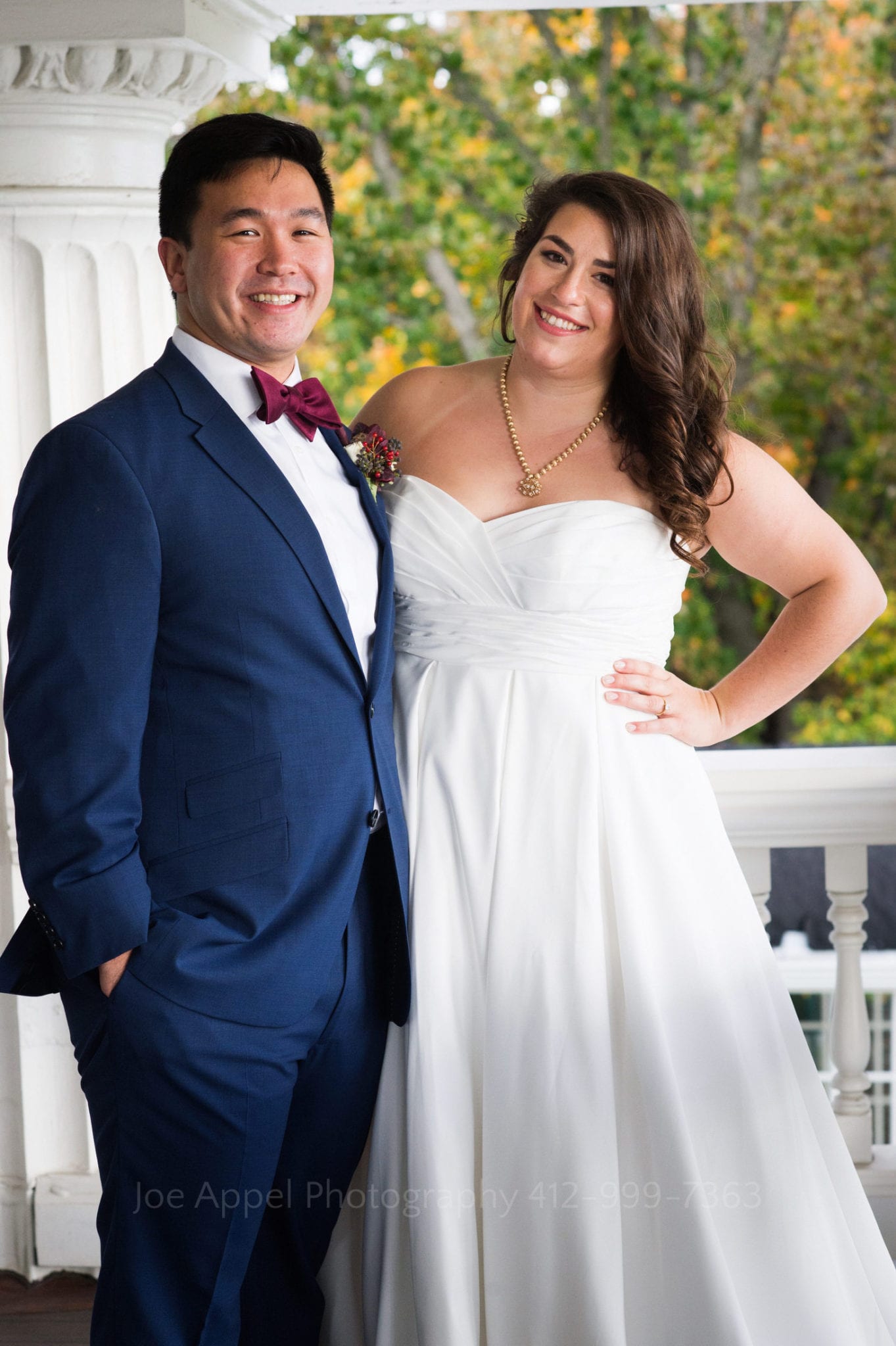 a groom in a blue suit stands on a white porch with his bride