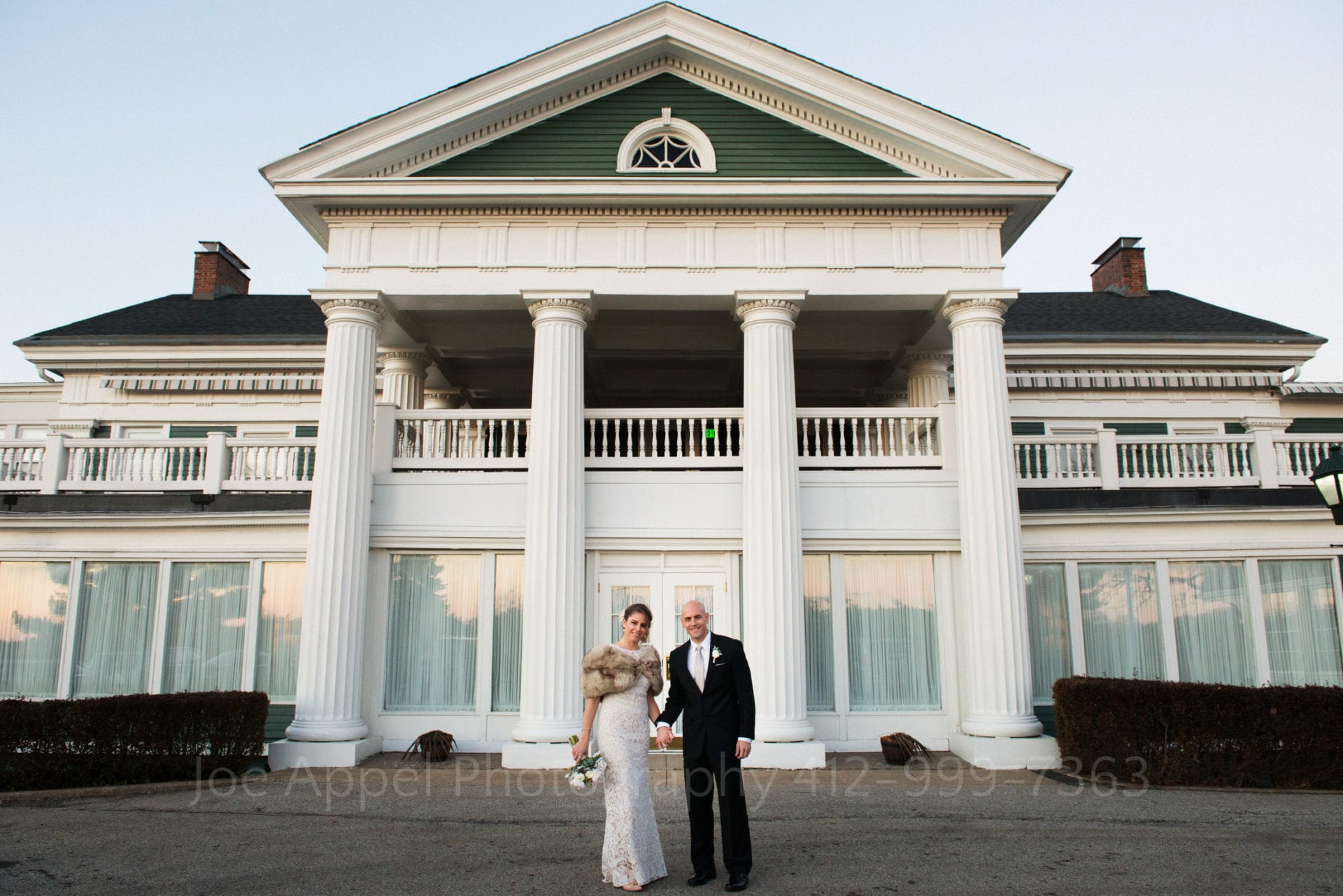 a bride in a fur parka stands with her groom in front of the pittsburgh golf club