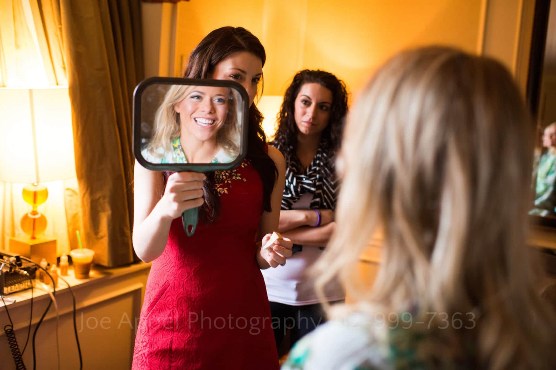 a bride smiles at her reflection in a hand mirror held by a bridesmaid