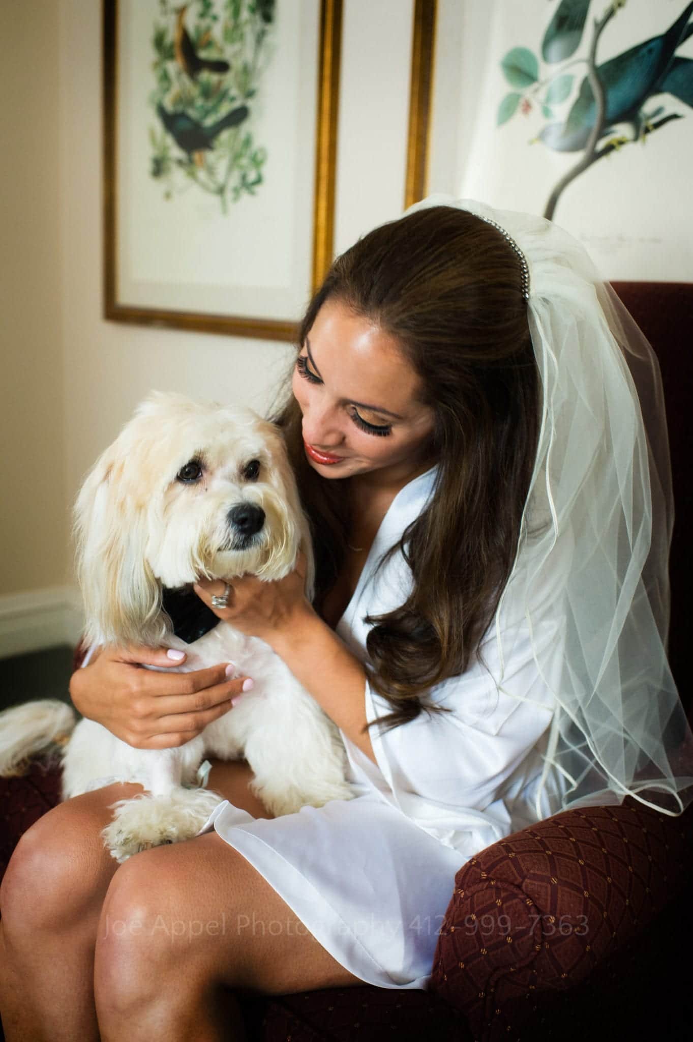 a brunette bride in a veil and silk white bathrobe pets a small white dog