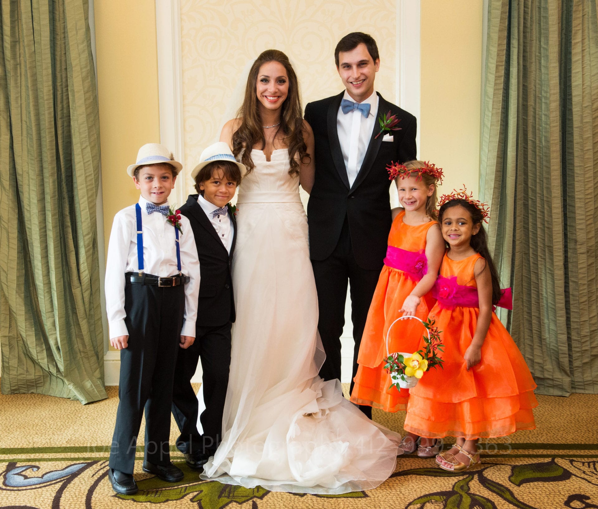 a bride and groom stand with their ring bearers in blue suspenders and flower girls in orange and pink dresses 