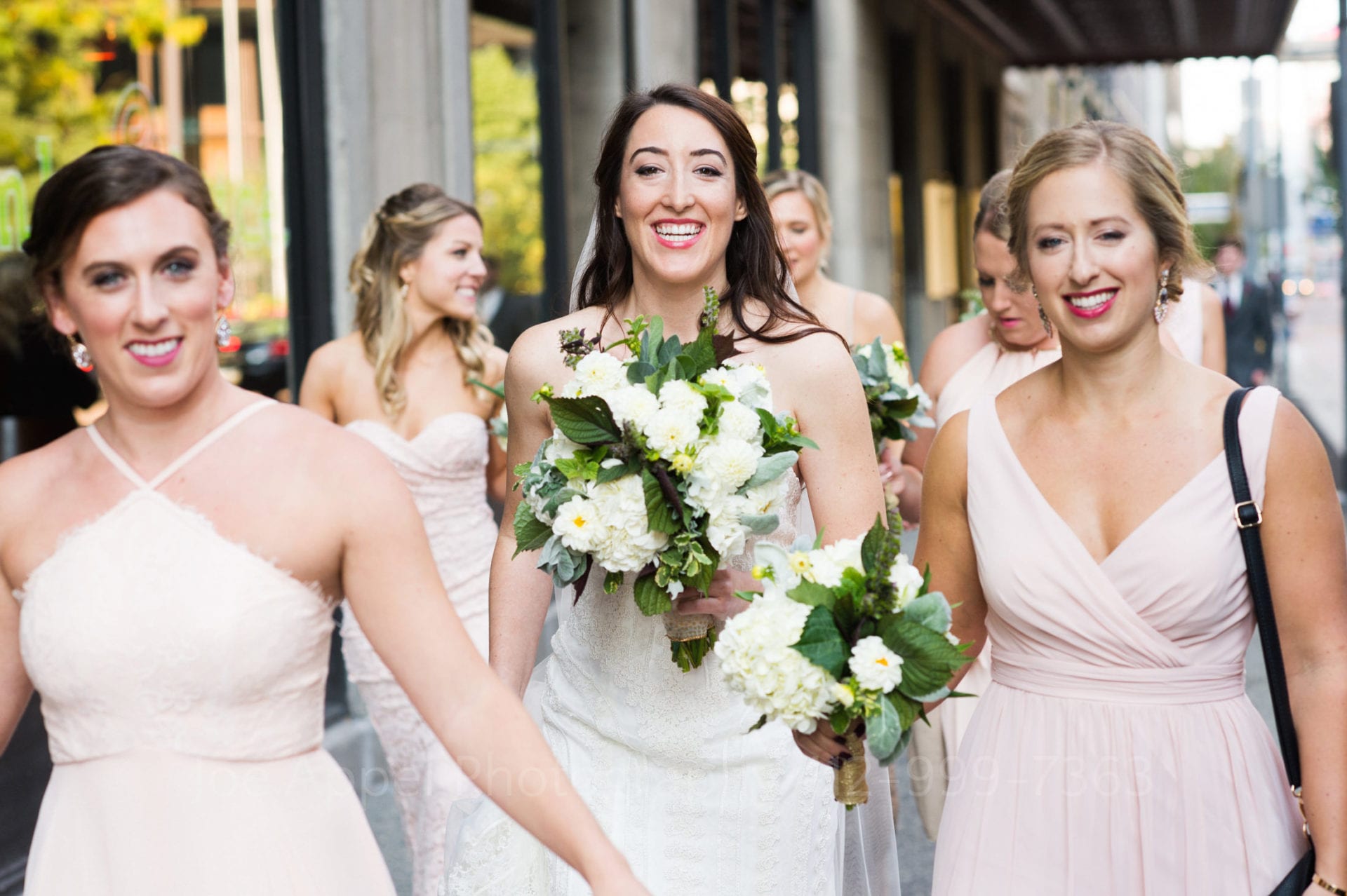 a bride holding a white bouquet walks with her bridesmaids wearing blush dresses