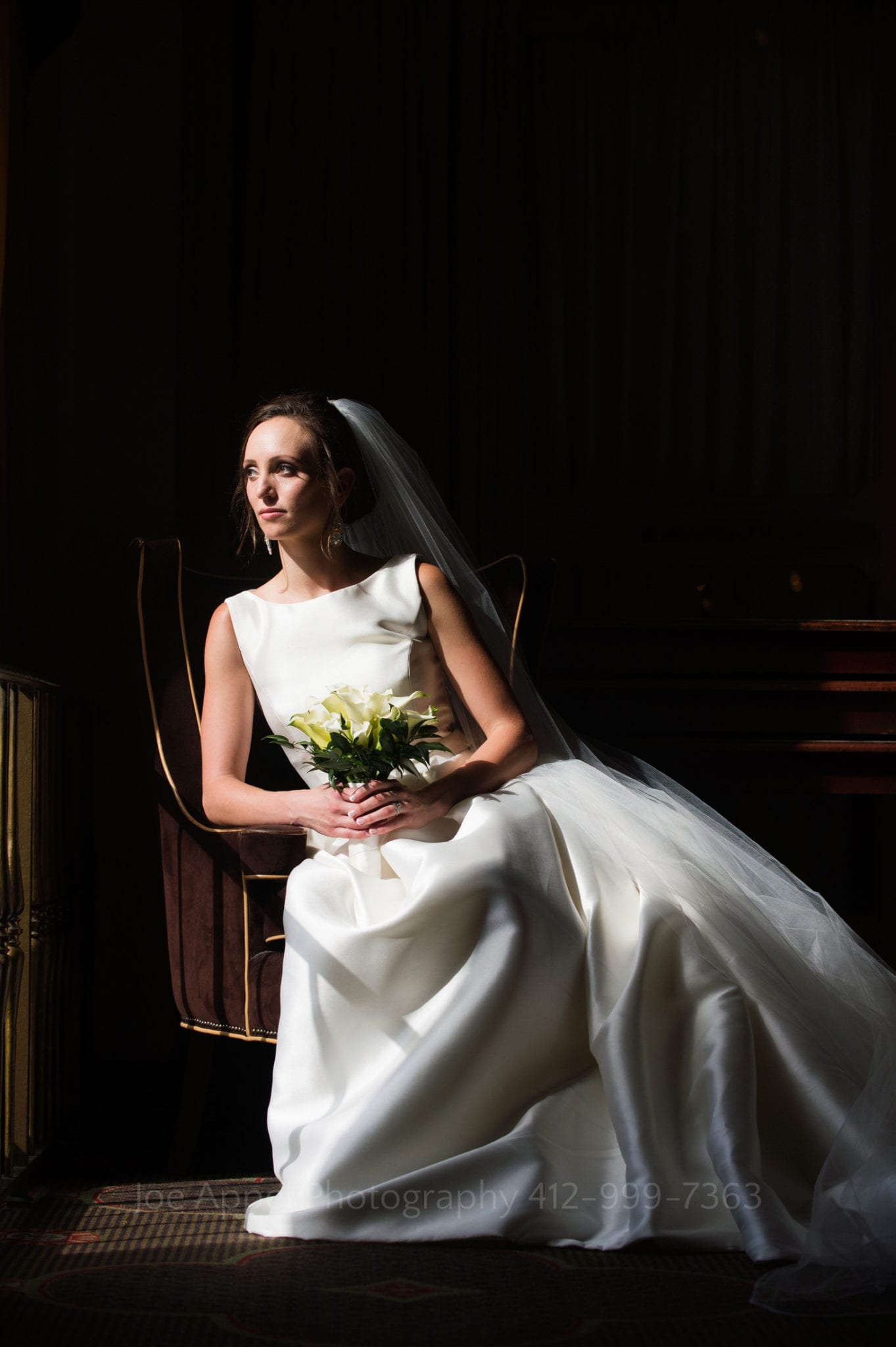 a bride with a white bouquet sits in a maroon arm chair and gazes out of the window