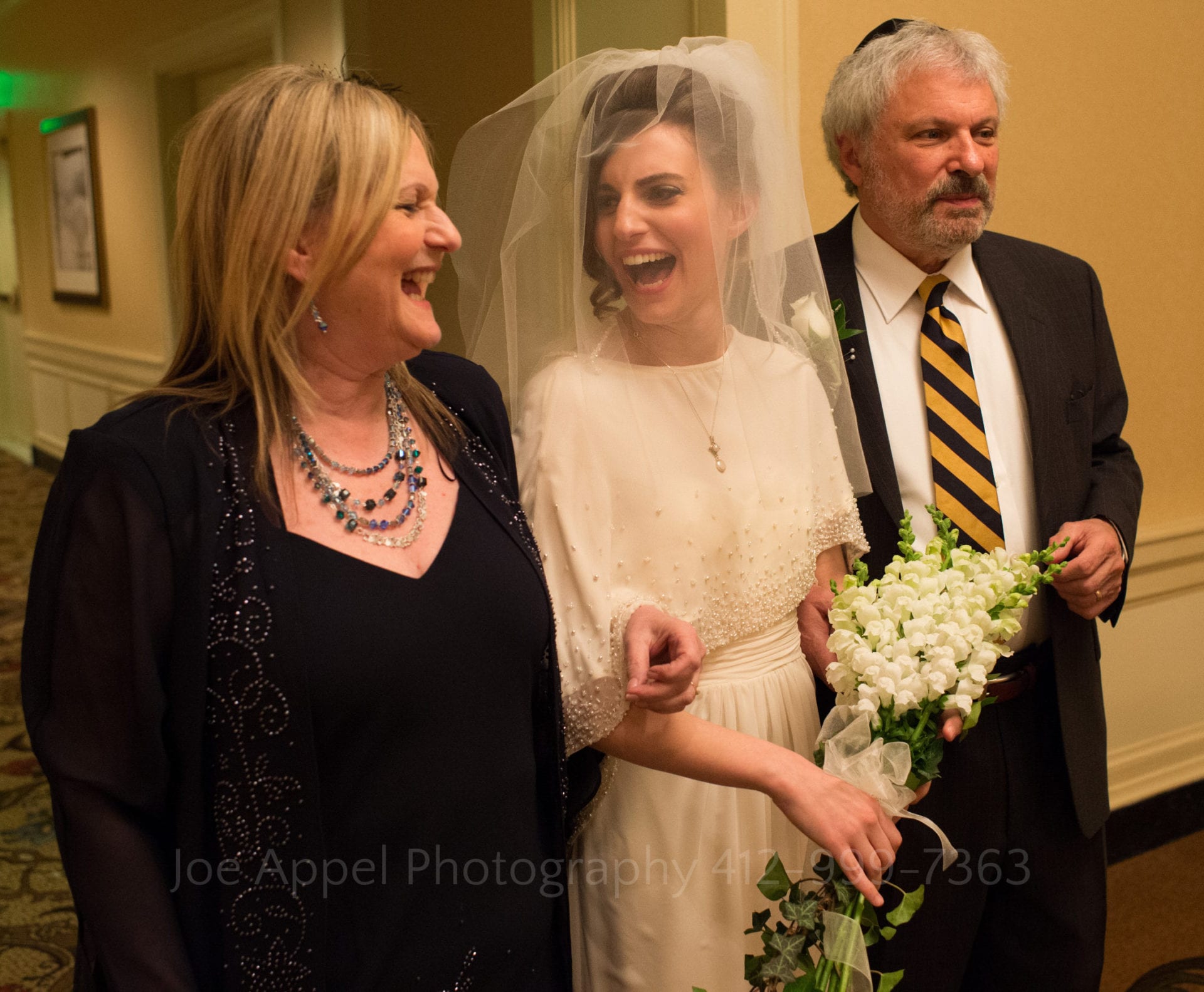 a bride in a white beaded dress holds a white bouquet and stands with her parents