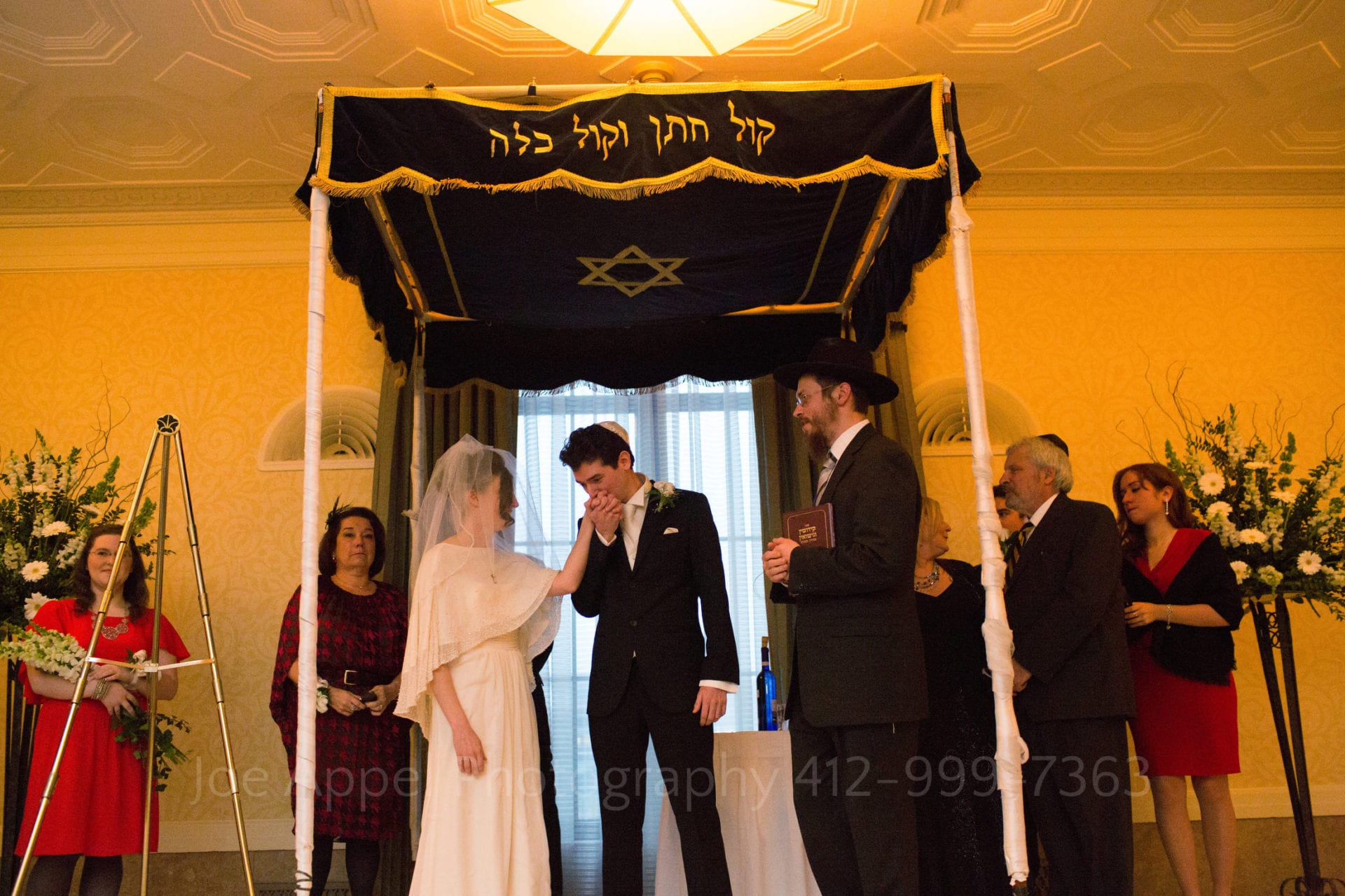 a groom kisses his brides hand as they stand under their chuppah