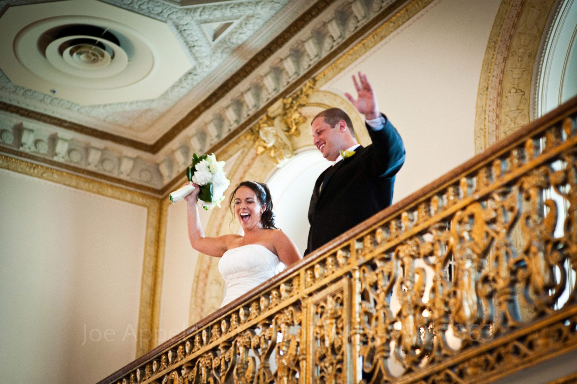 a bride and groom wave at their guests from a gold balcony