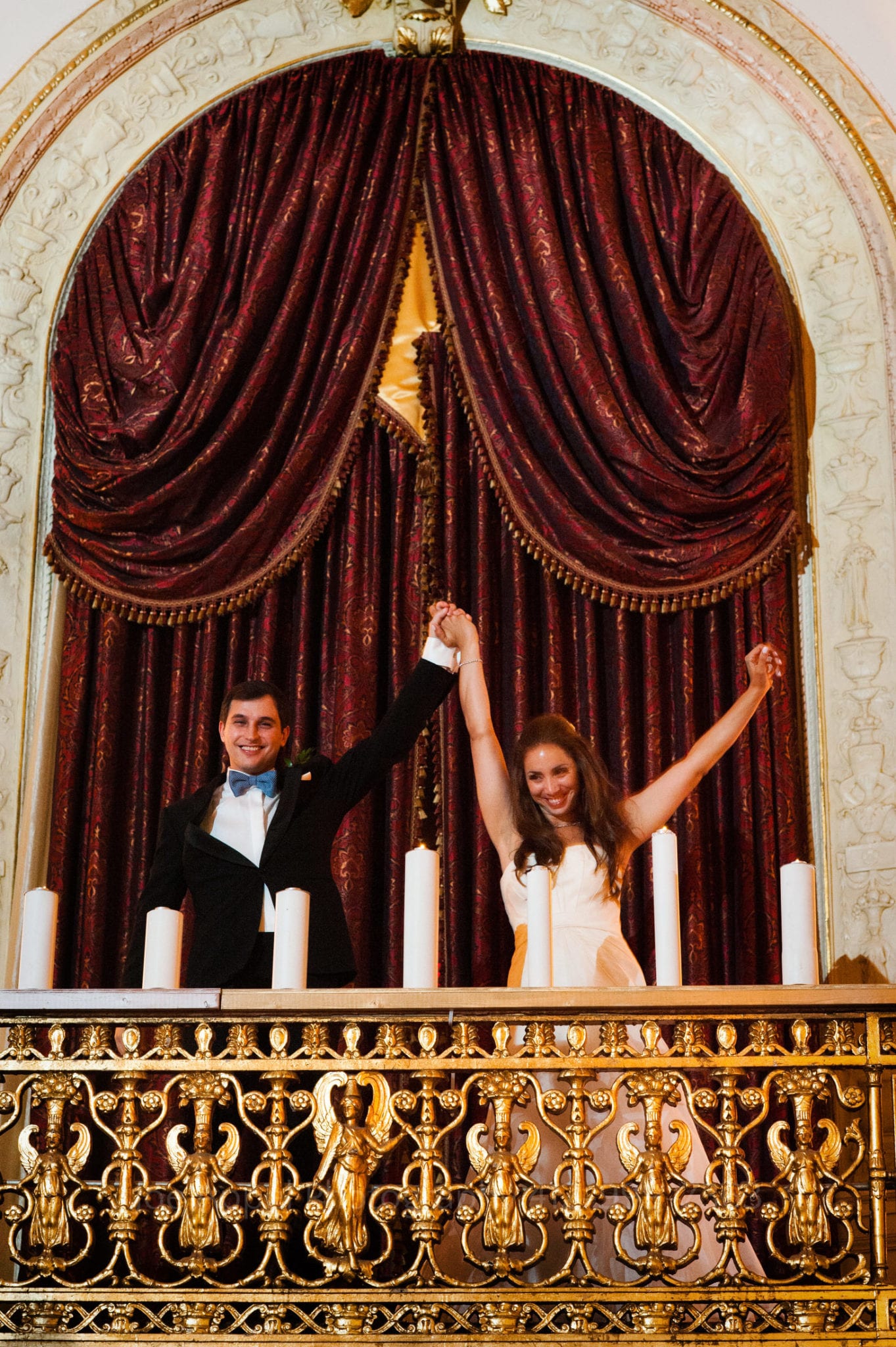 a bride and groom stand in front of a red curtain on a gold balcony
