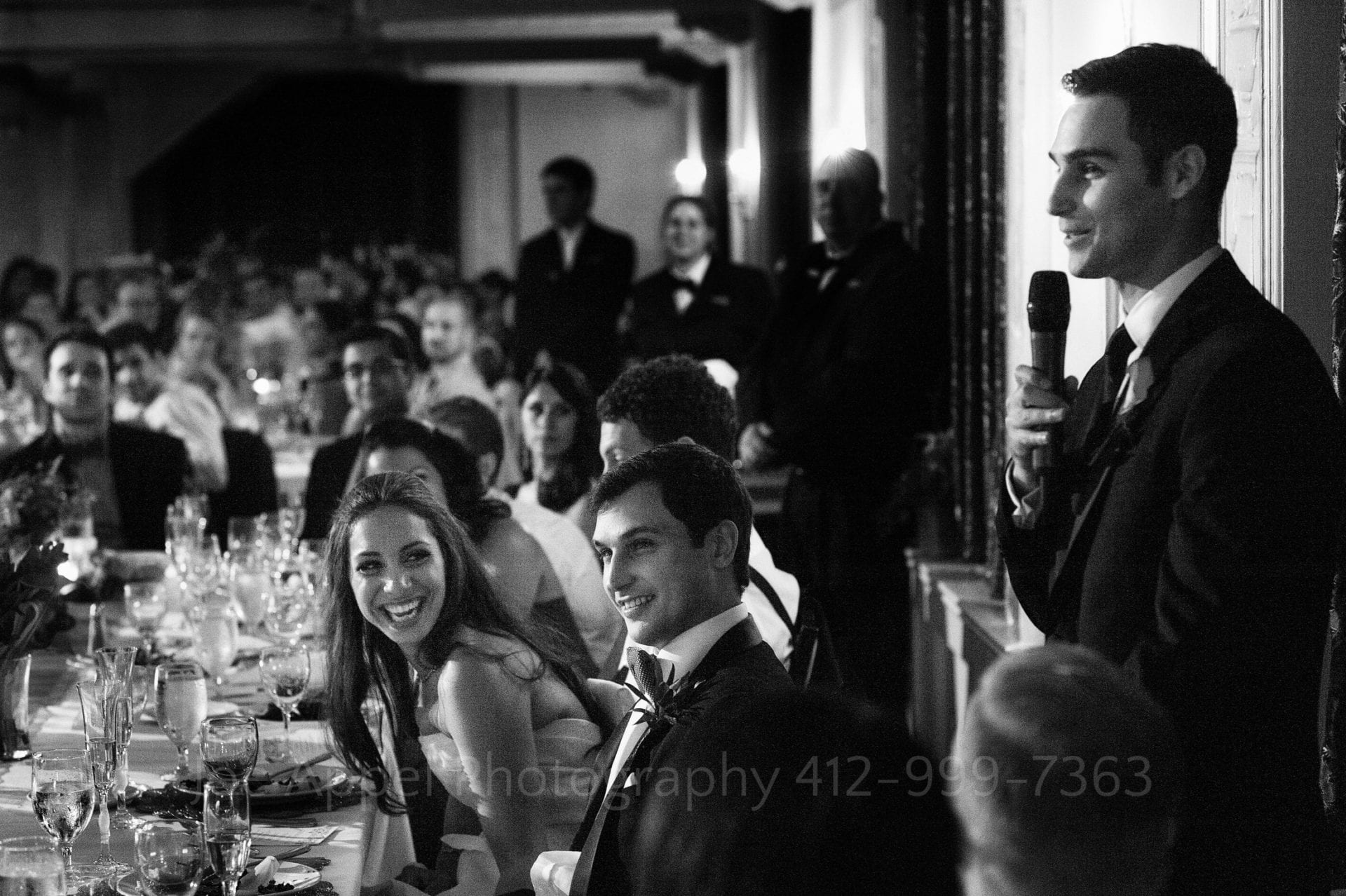 a bride and groom laugh as their friends make speeches