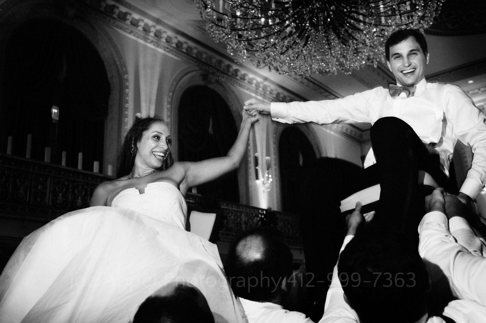 a bride and groom laugh as their guests lift them in chairs