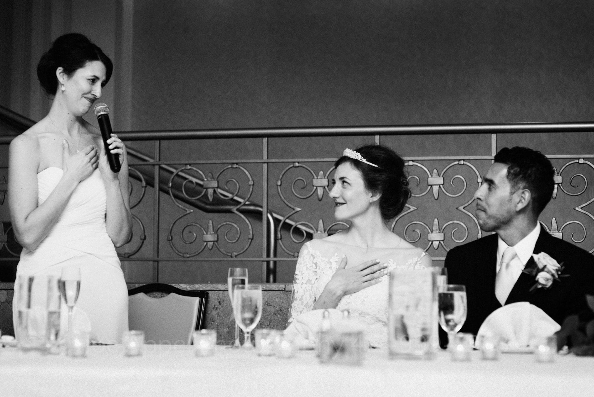 a bride and groom smile as their friend makes a speech