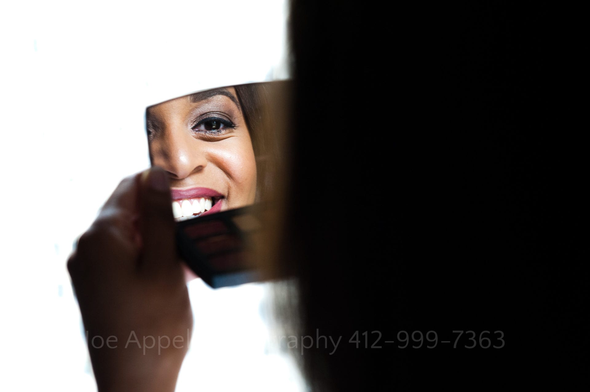 A young woman's eye and smile are reflected in a small mirror in front of a window at an Omni William Penn weddings