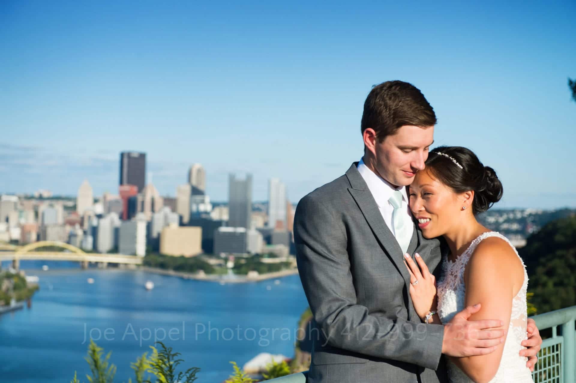 a bride and groom stand with a blue sky and a view of downtown pittsburgh behind them