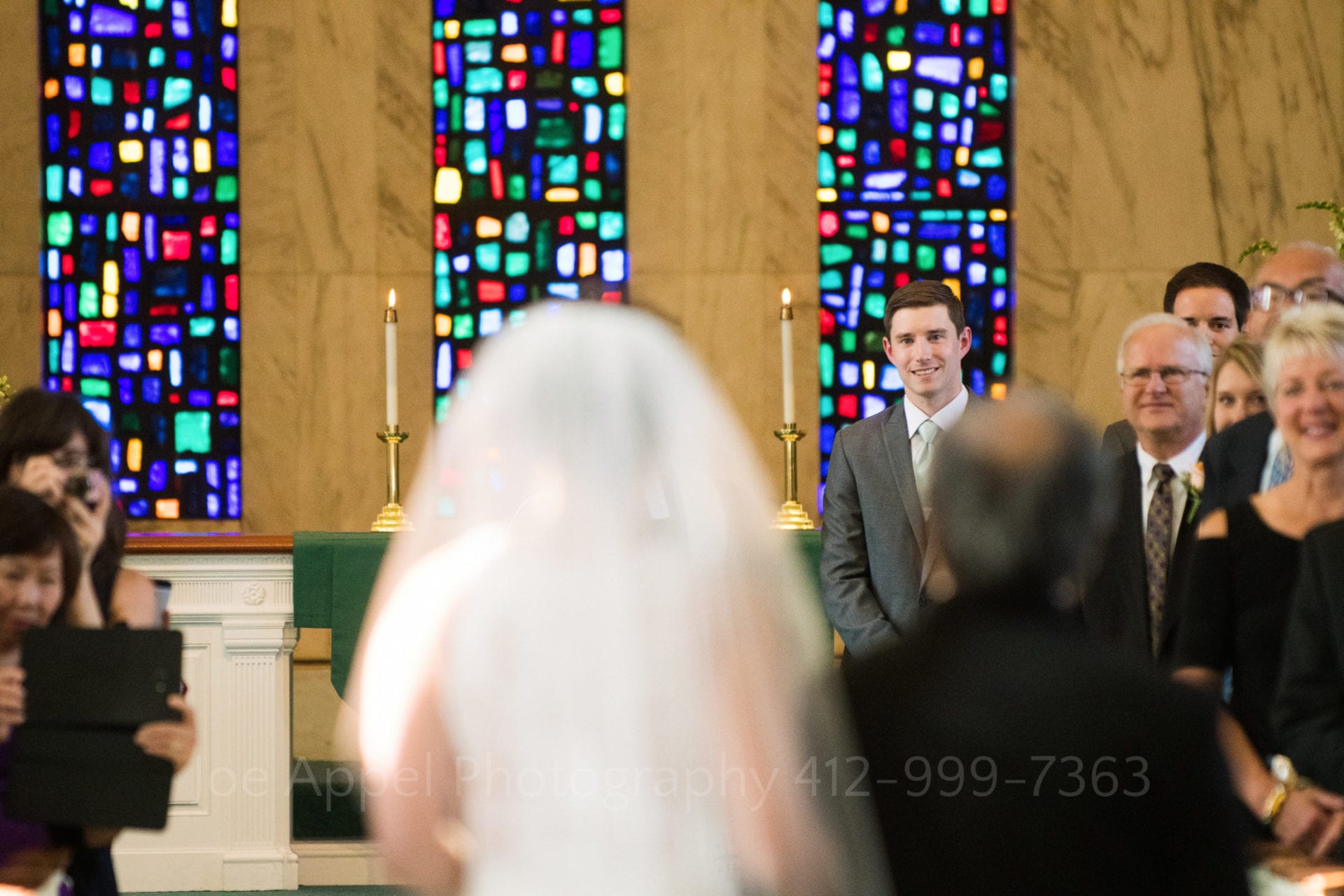 a groom stands in front of colorful stained glass windows and smiles at his bride