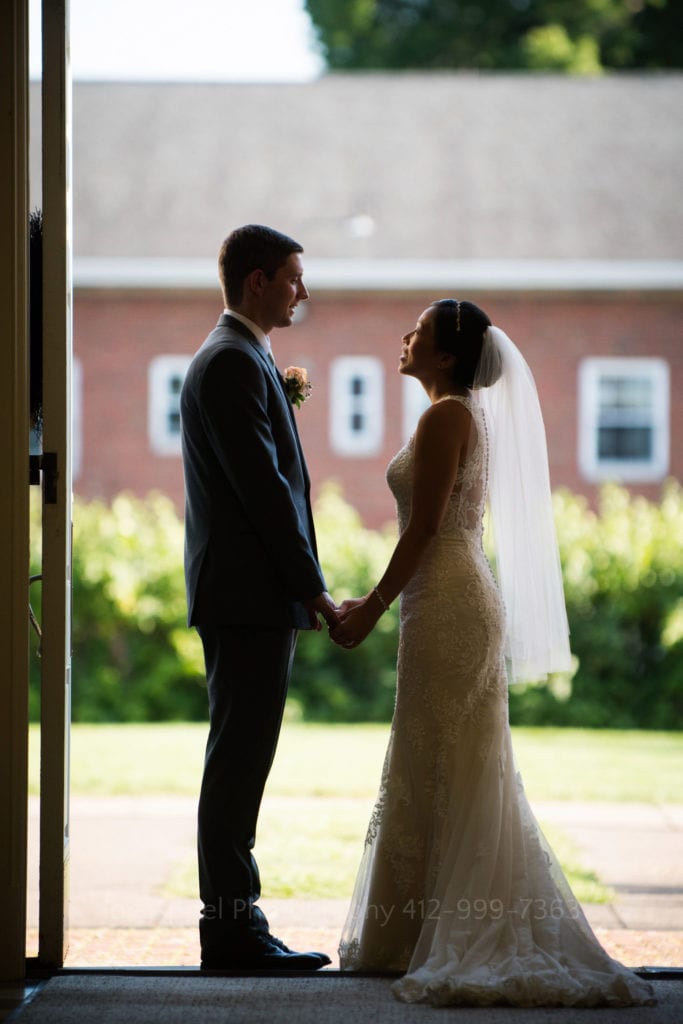 a bride and groom hold hands and talk in a sunlit doorway