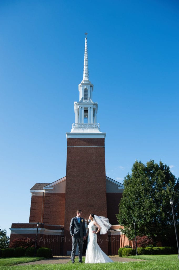 a bride and groom pose in front of a chapel with a blue sky behind them
