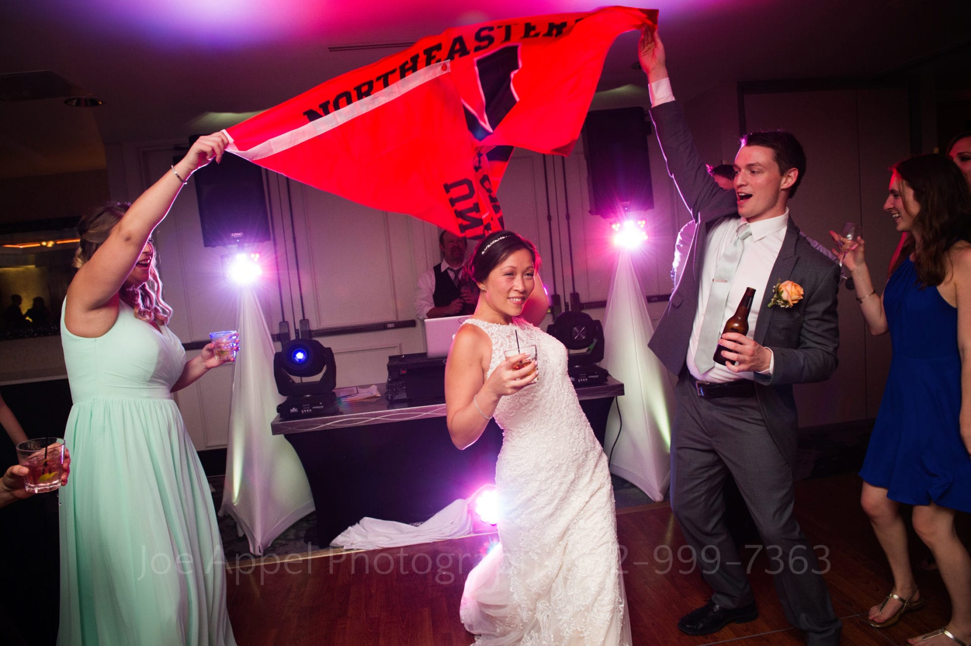 a bridesmaid and groomsman wave a red northeastern university flag above a bride