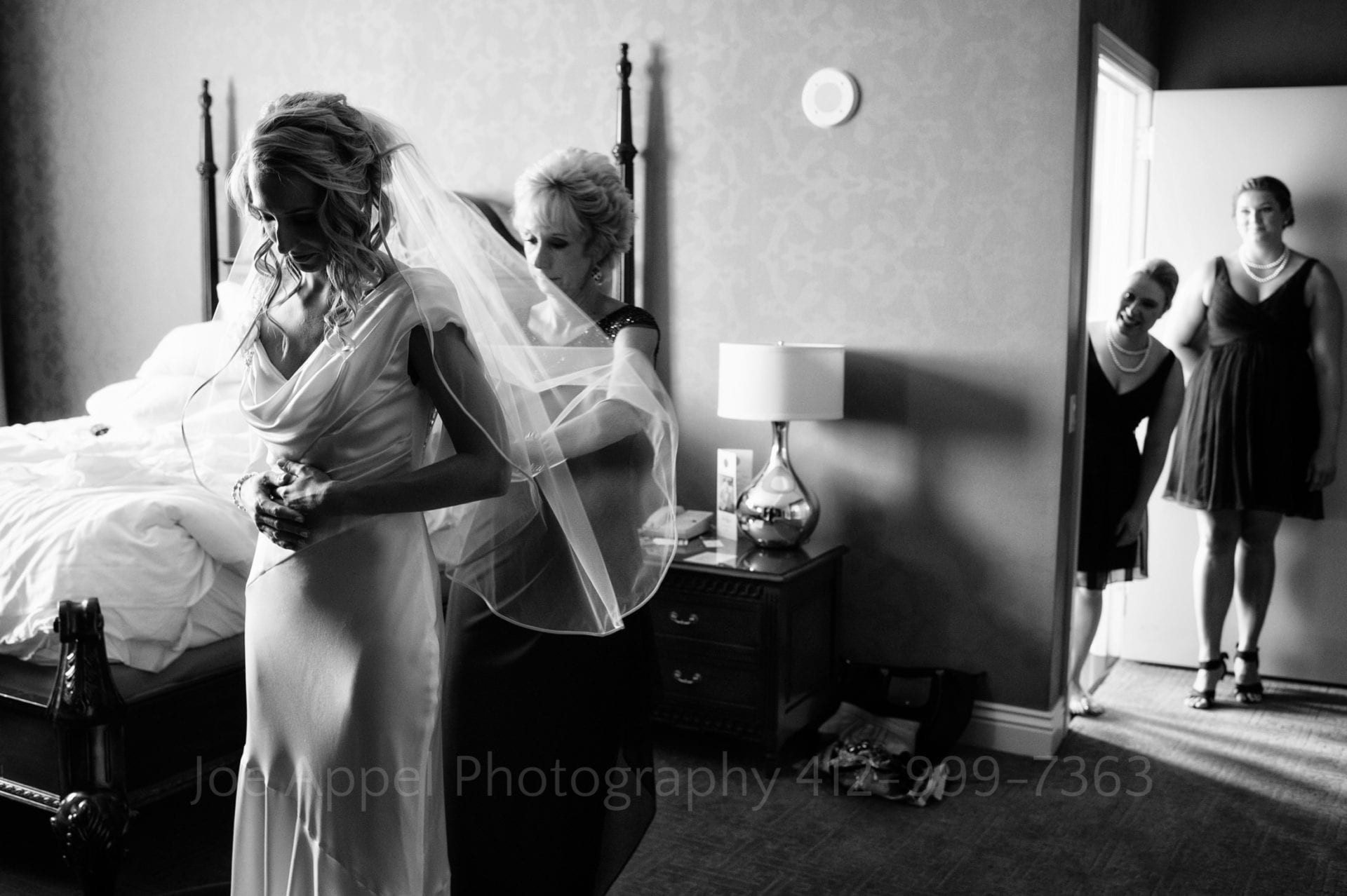 two bridesmaids smile and watch a brides mother button her wedding dress