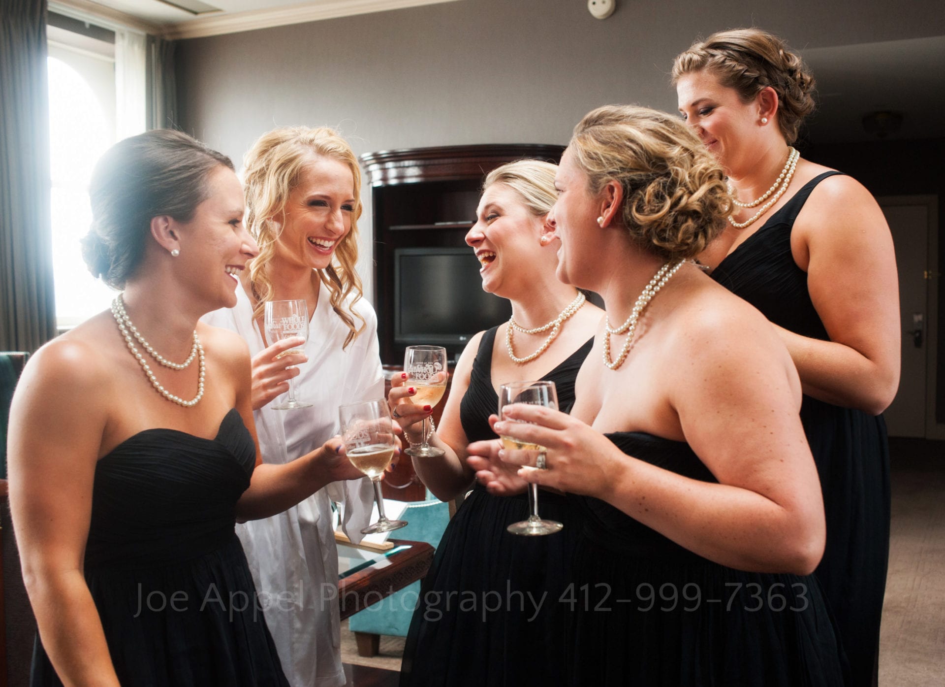 a bride laughs and toasts champagne with bridesmaids wearing black dresses and pearls