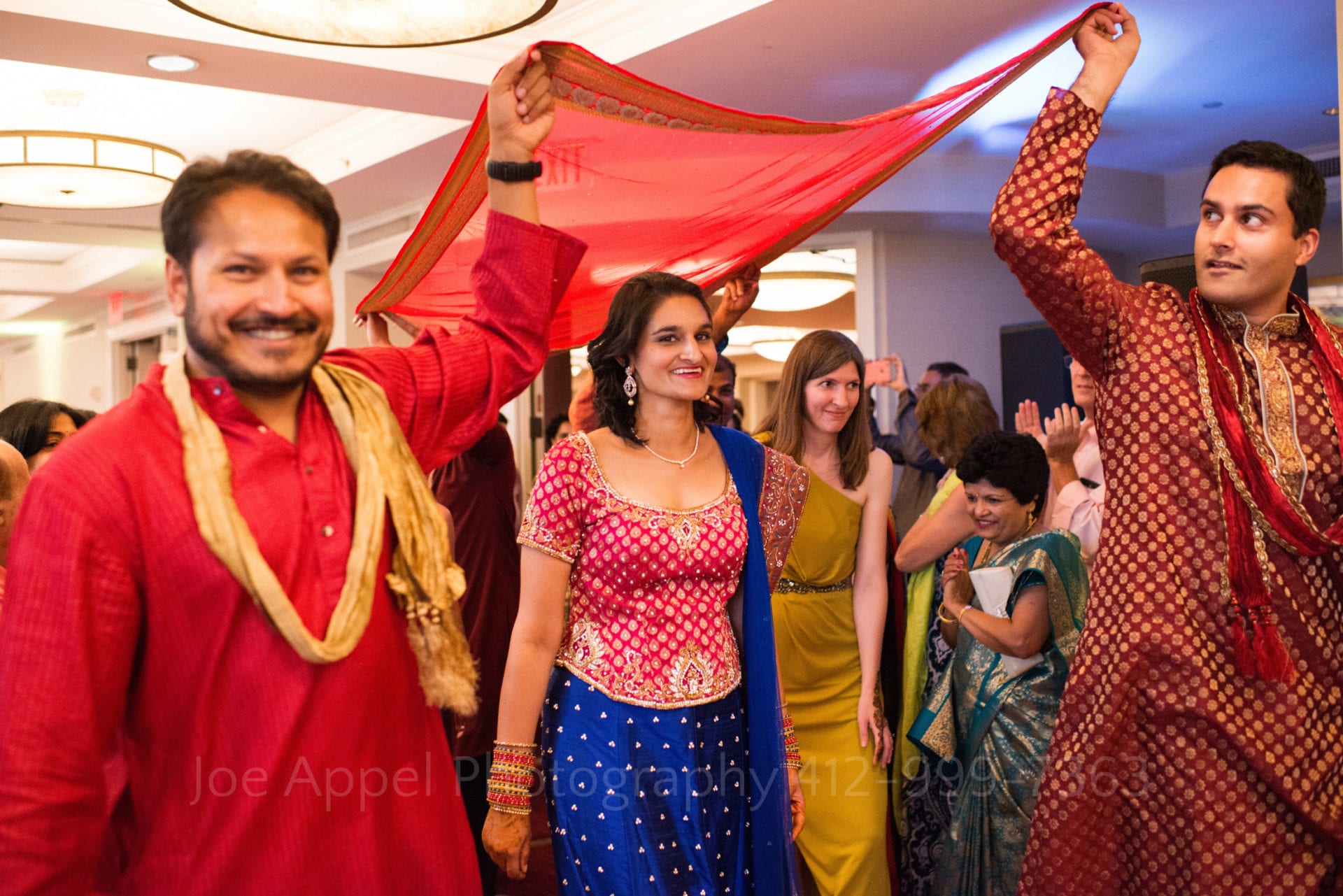 men in red kurtas hold a red and gold sheet over a bride wearing a red and blue sari